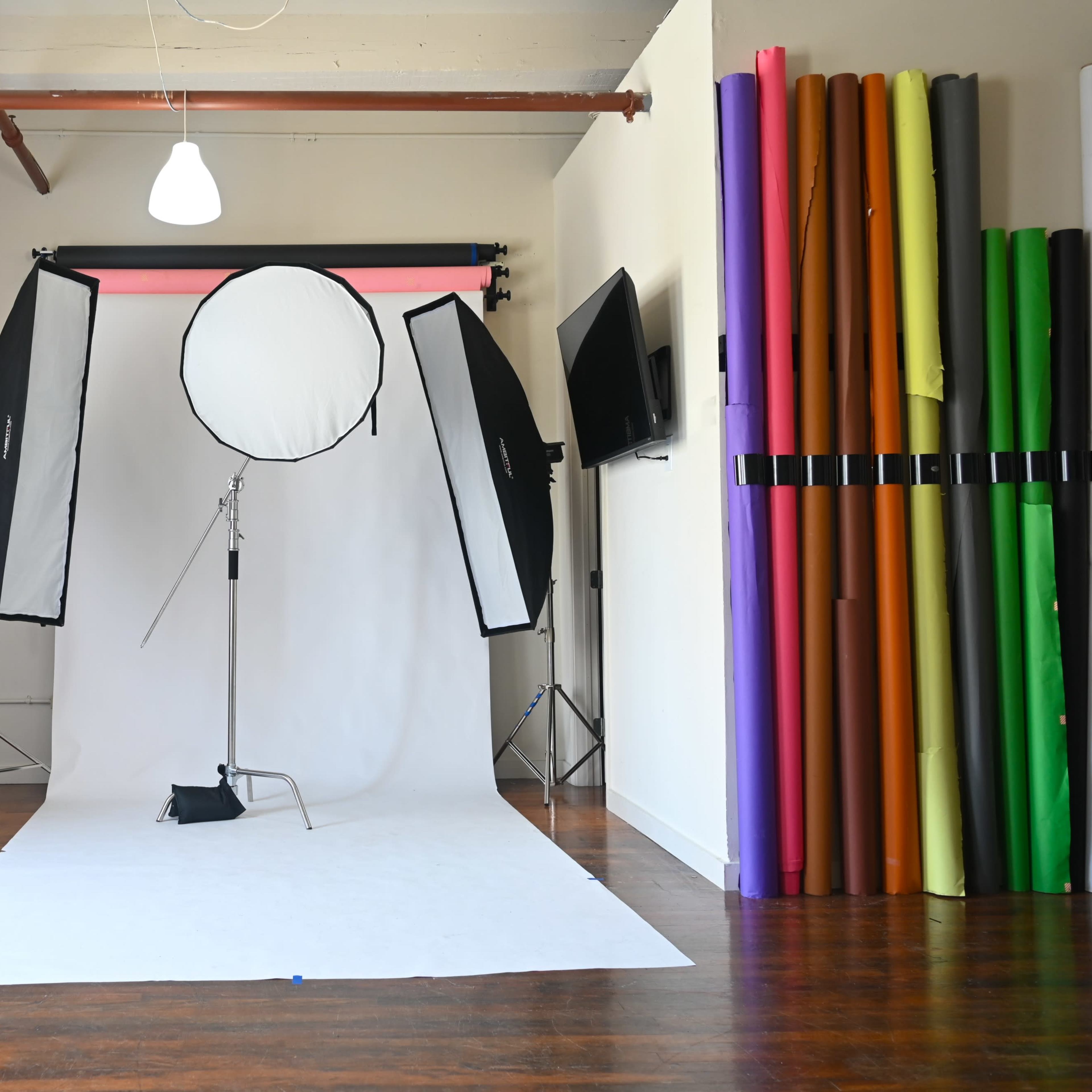 A photography studio with softbox lighting setups, a white backdrop, and a collection of colorful backdrop rolls on one side.