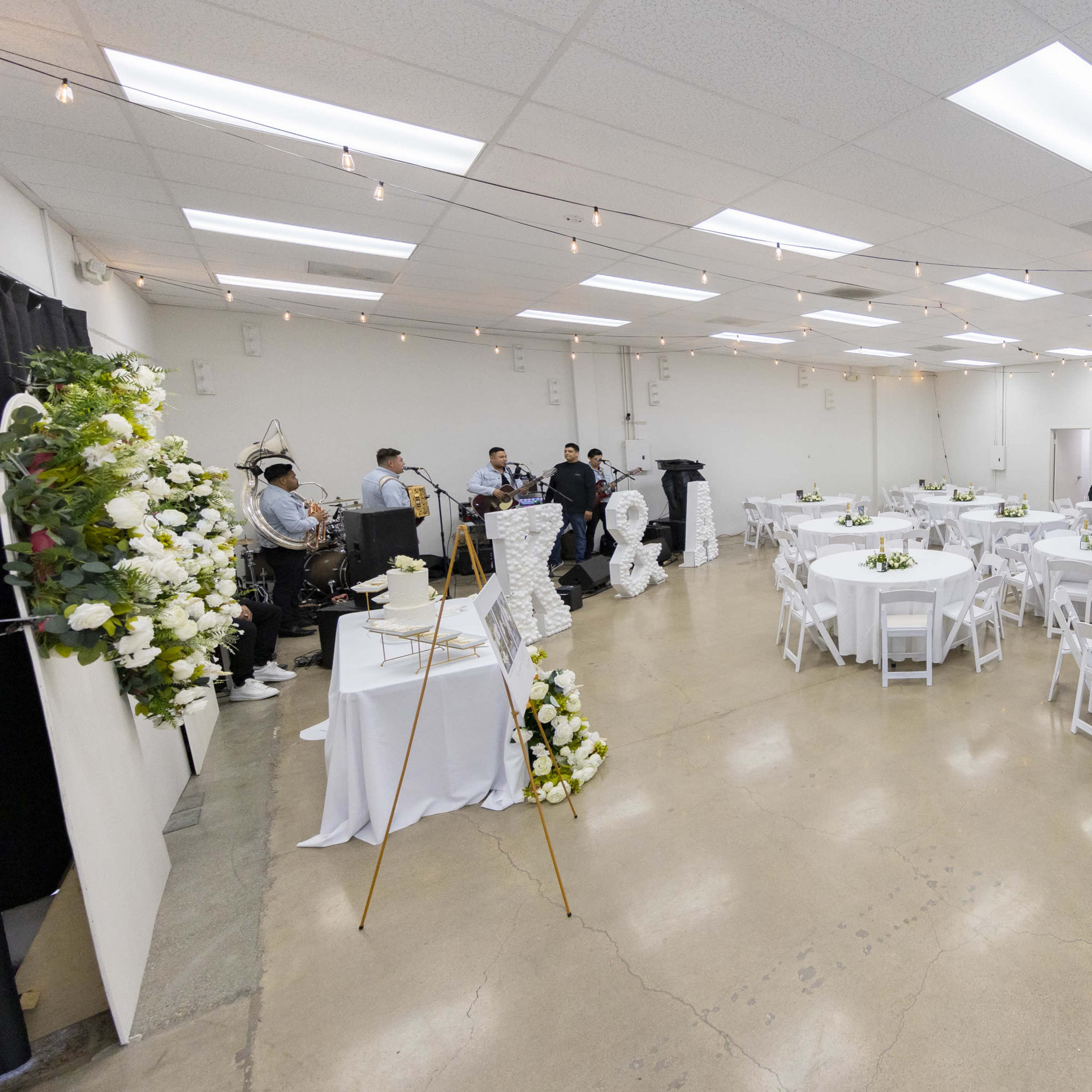 A spacious event hall set up for a gathering, with round tables decorated with flowers and a small buffet area on one side.