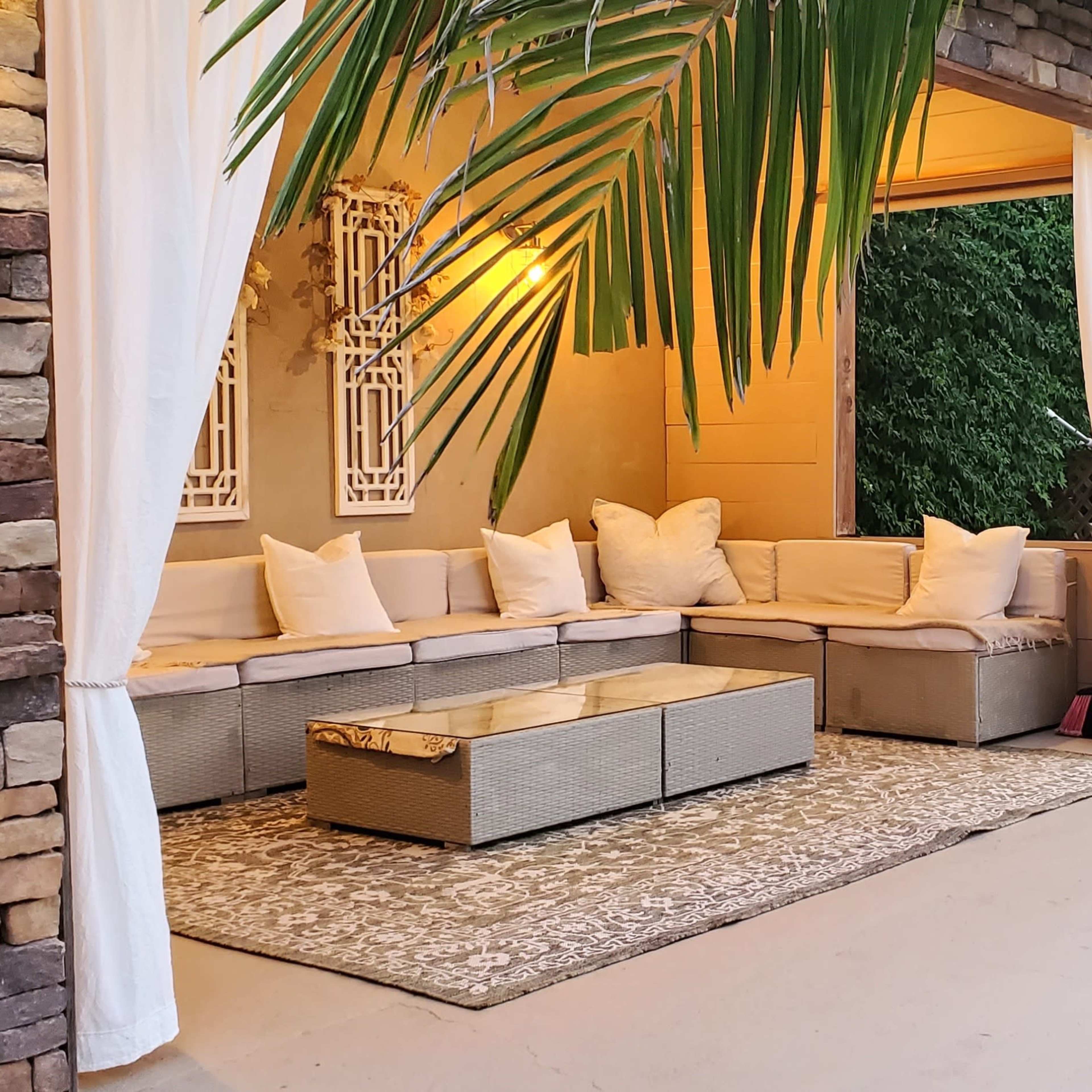 A landscaped outdoor seating area features a sectional sofa with cushions, a coffee table, and decorative elements surrounded by palm leaves and stone walls.