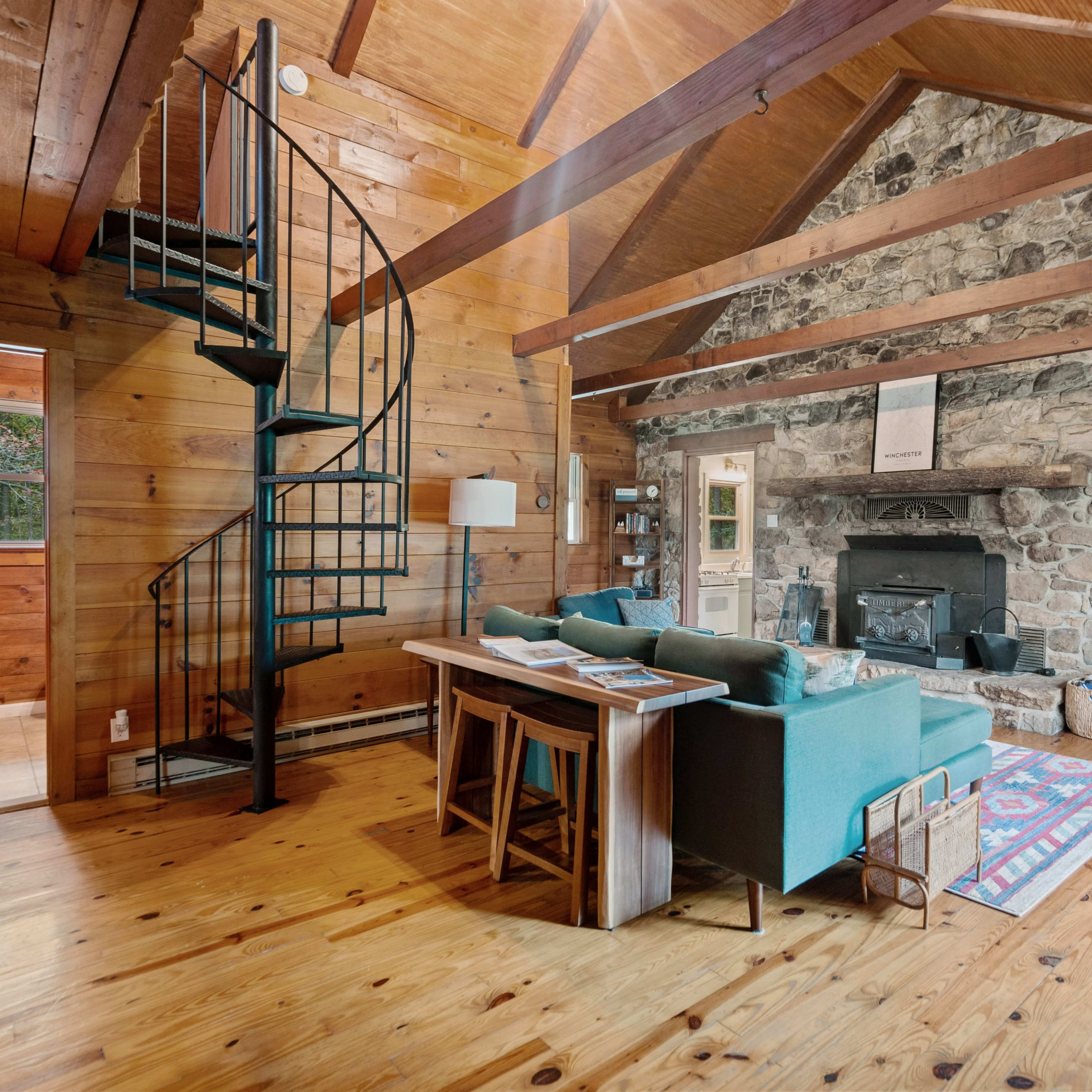 A cozy living area with a stone fireplace, wooden beams, a spiral staircase, and a turquoise sofa facing a television.