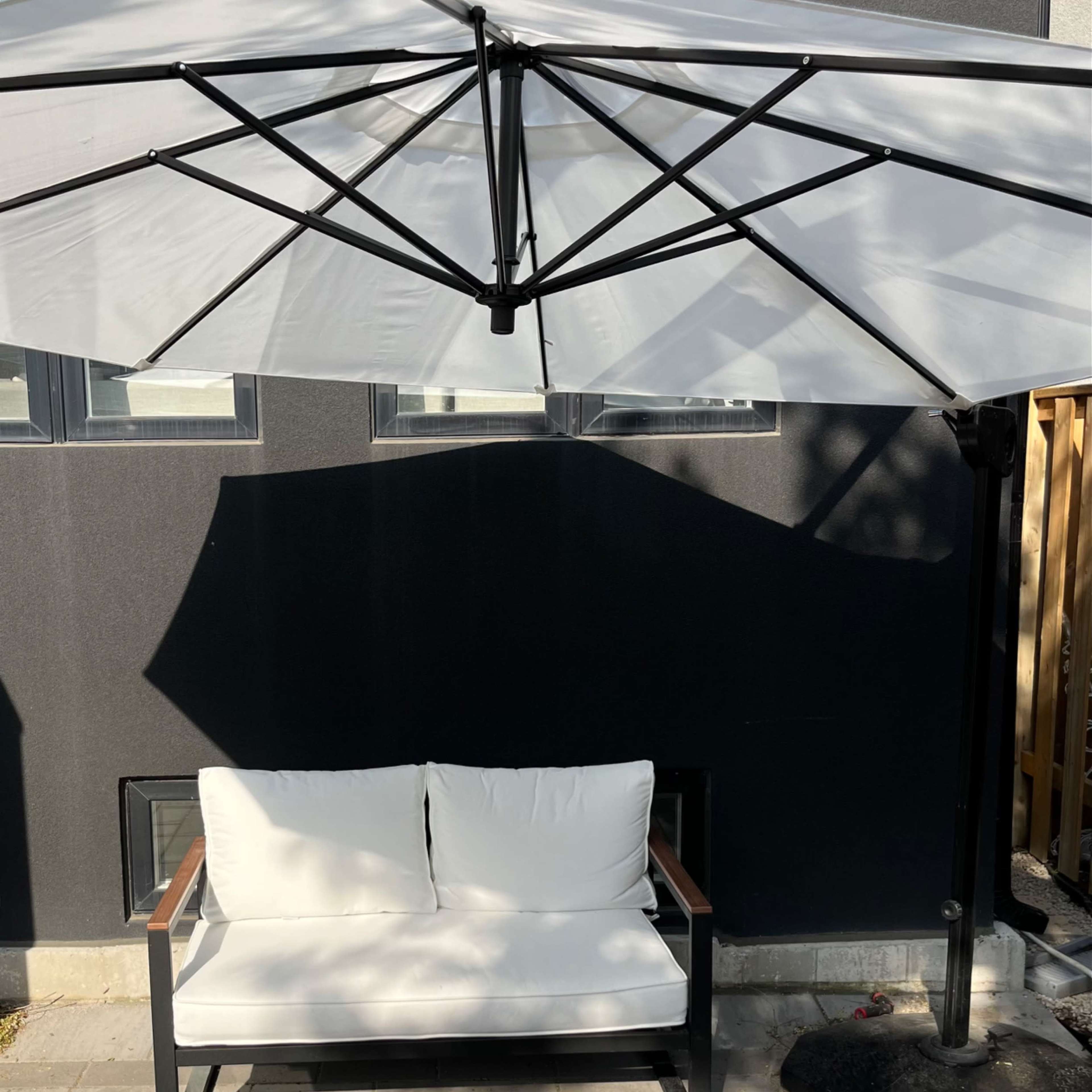 A white couch with a wooden armrest sits under a large umbrella on a stone patio.