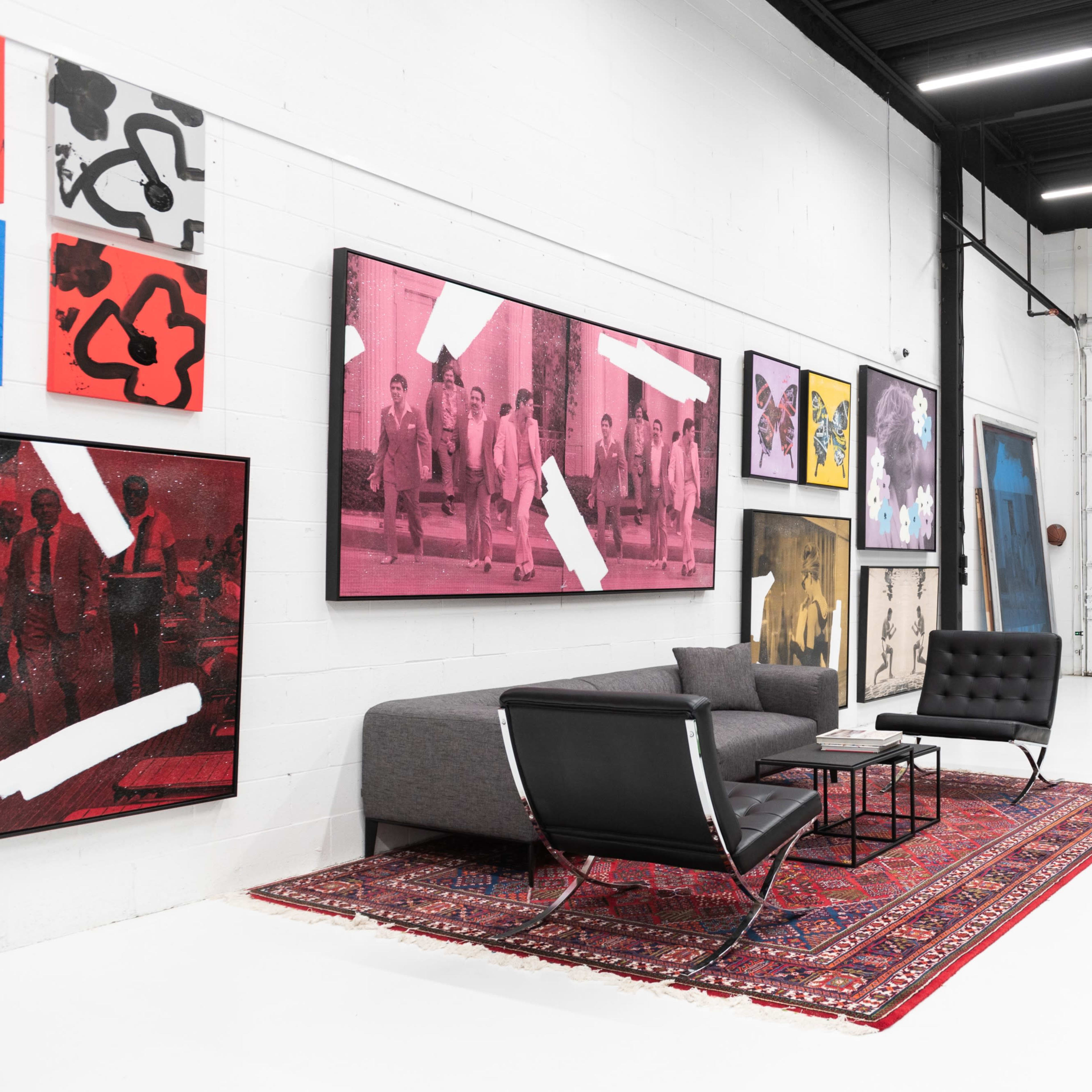 A modern gallery space featuring a gray sofa set beside a colorful collection of framed artworks on the white walls, with a patterned rug on the floor.