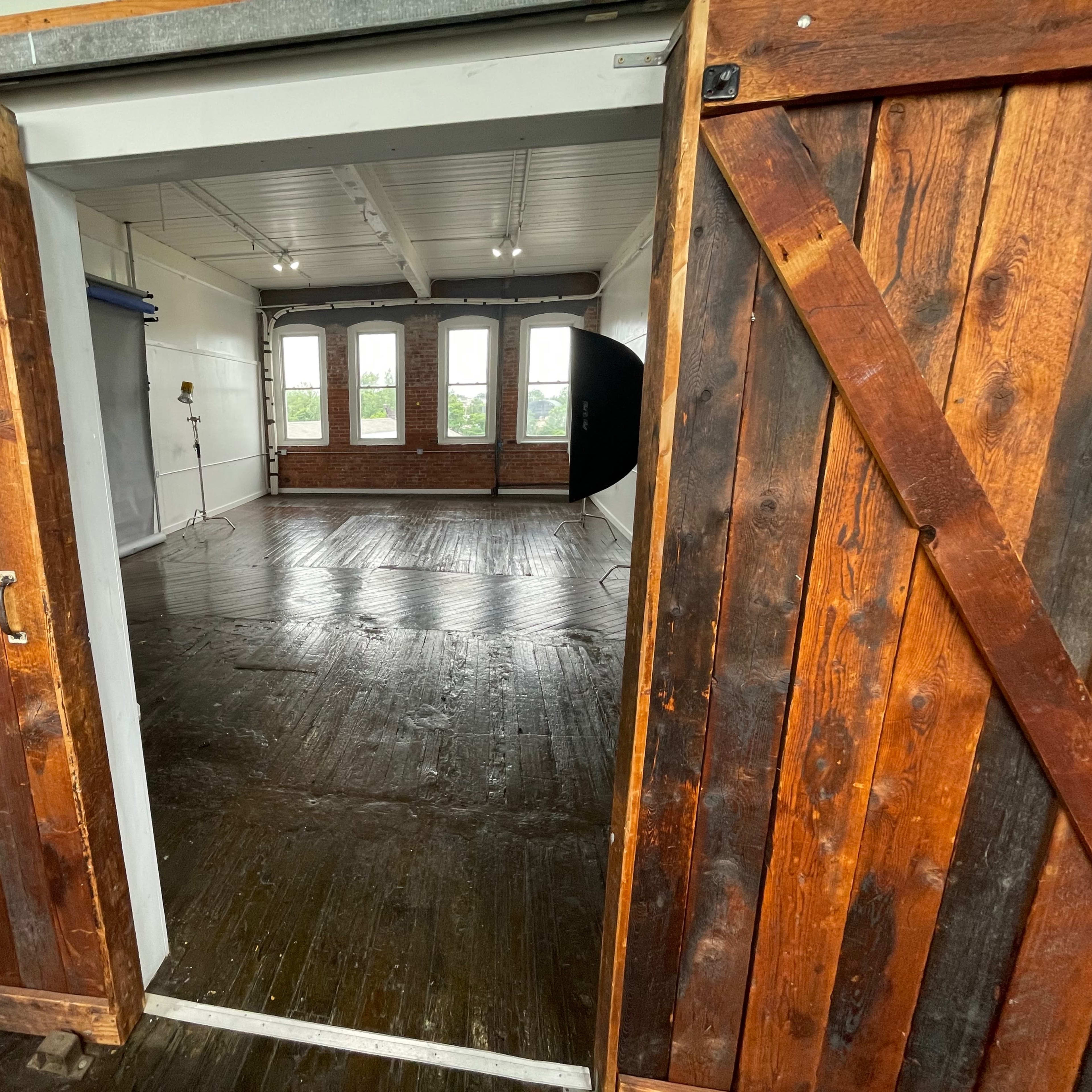 An open wooden double door leading into a bright room with large windows and wooden floors.