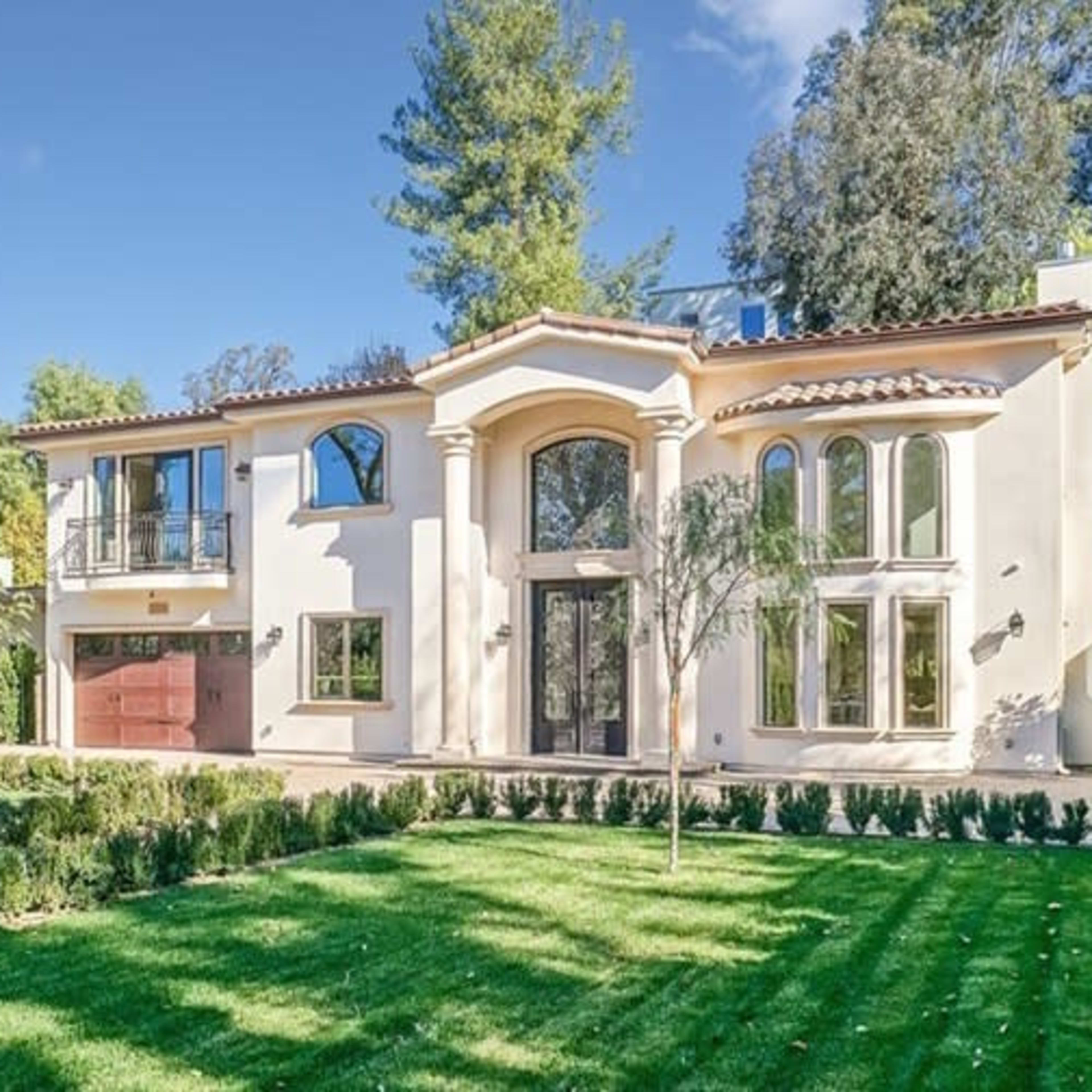 A large, two-story Mediterranean-style house with a red garage door and landscaped front yard is surrounded by trees and shrubs.