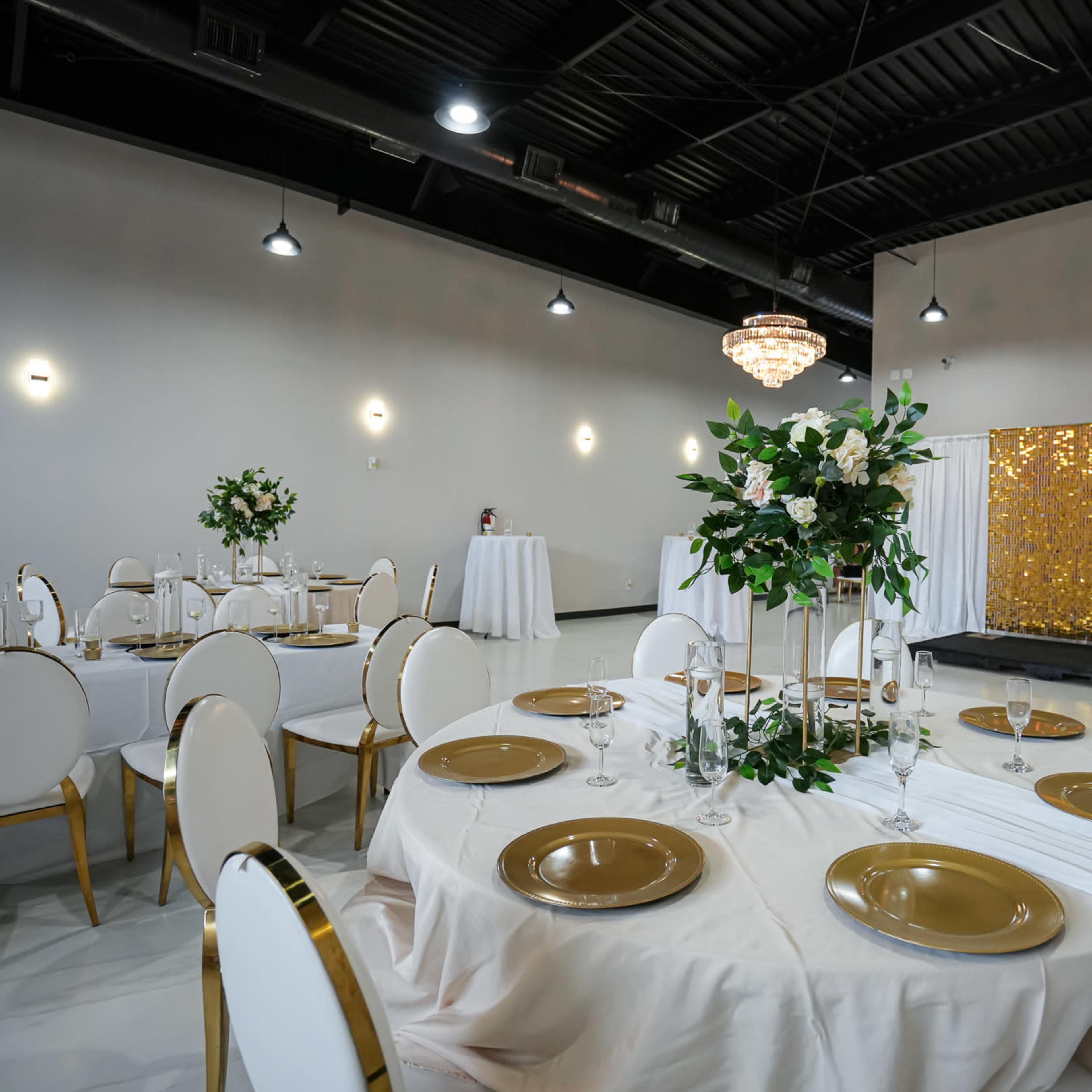 A dining area is set up with round tables adorned in white linens and gold accents, featuring floral centerpieces, with a backdrop of a glittering gold curtain.