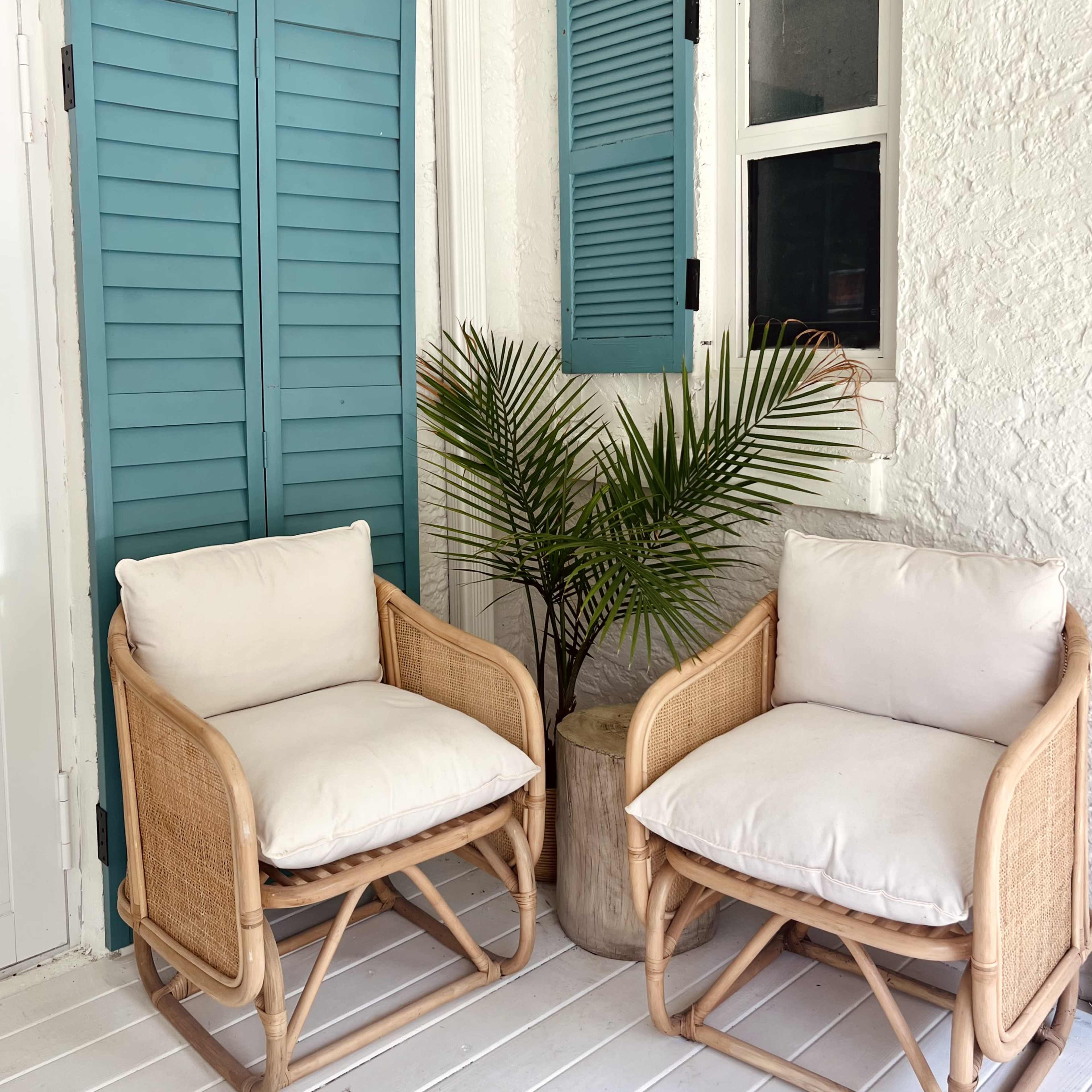 Two rattan chairs with white cushions are positioned beside a potted palm plant against a backdrop of blue shutters and a white wall.