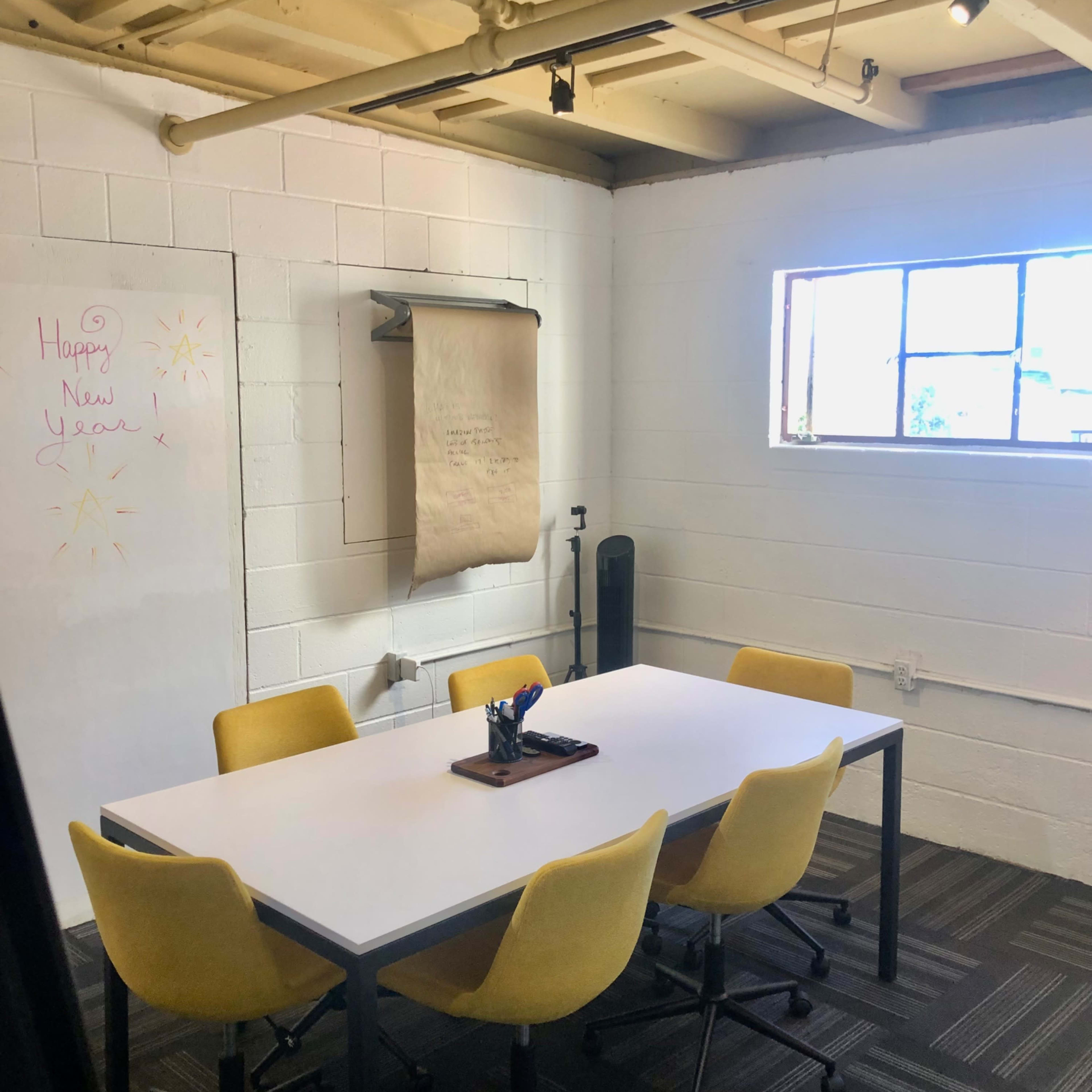 A conference room features a white table surrounded by yellow chairs, with a window on one wall and a large paper roll mounted for notes.