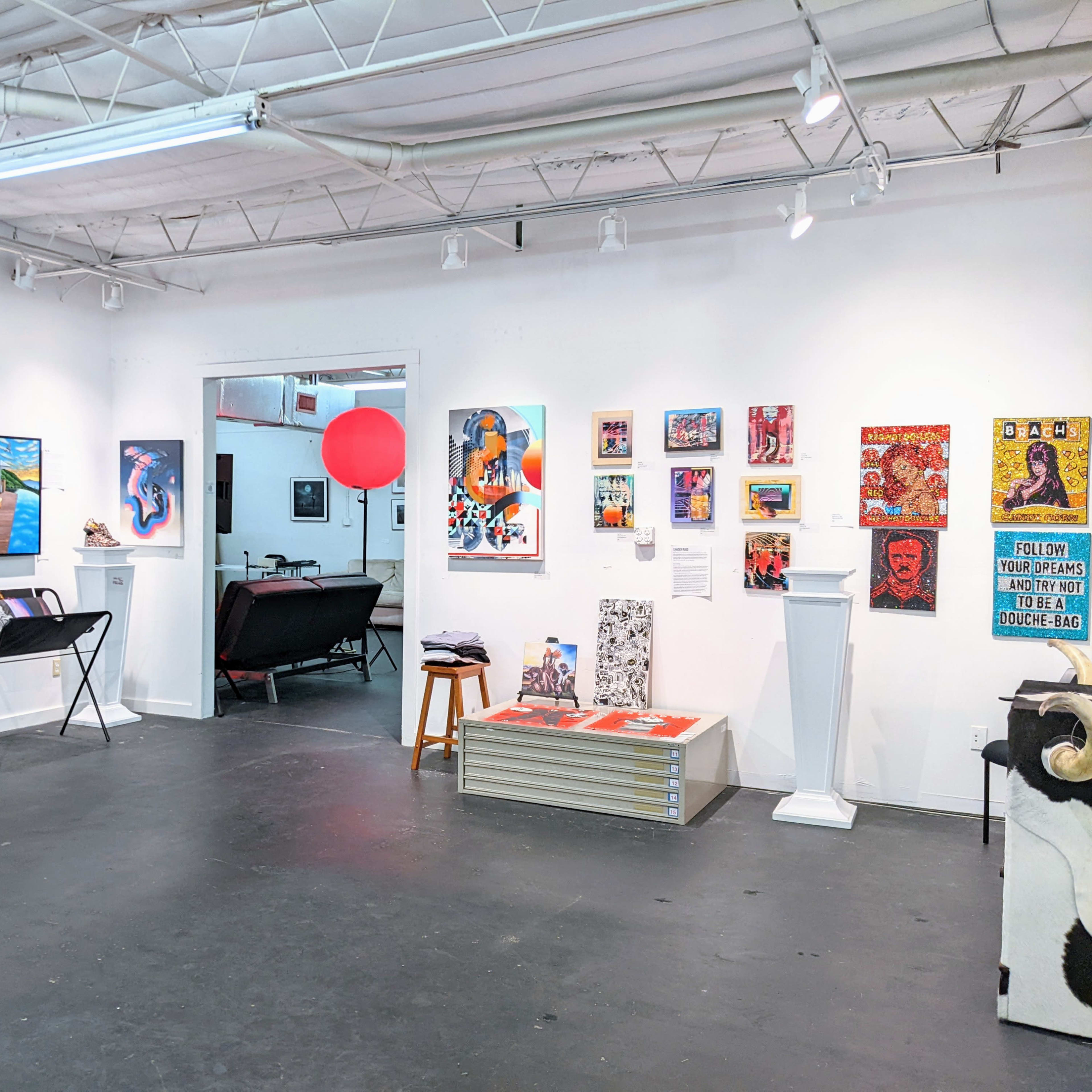 An art gallery interior featuring colorful artwork on white walls, clothing displayed on racks, and various furniture pieces.