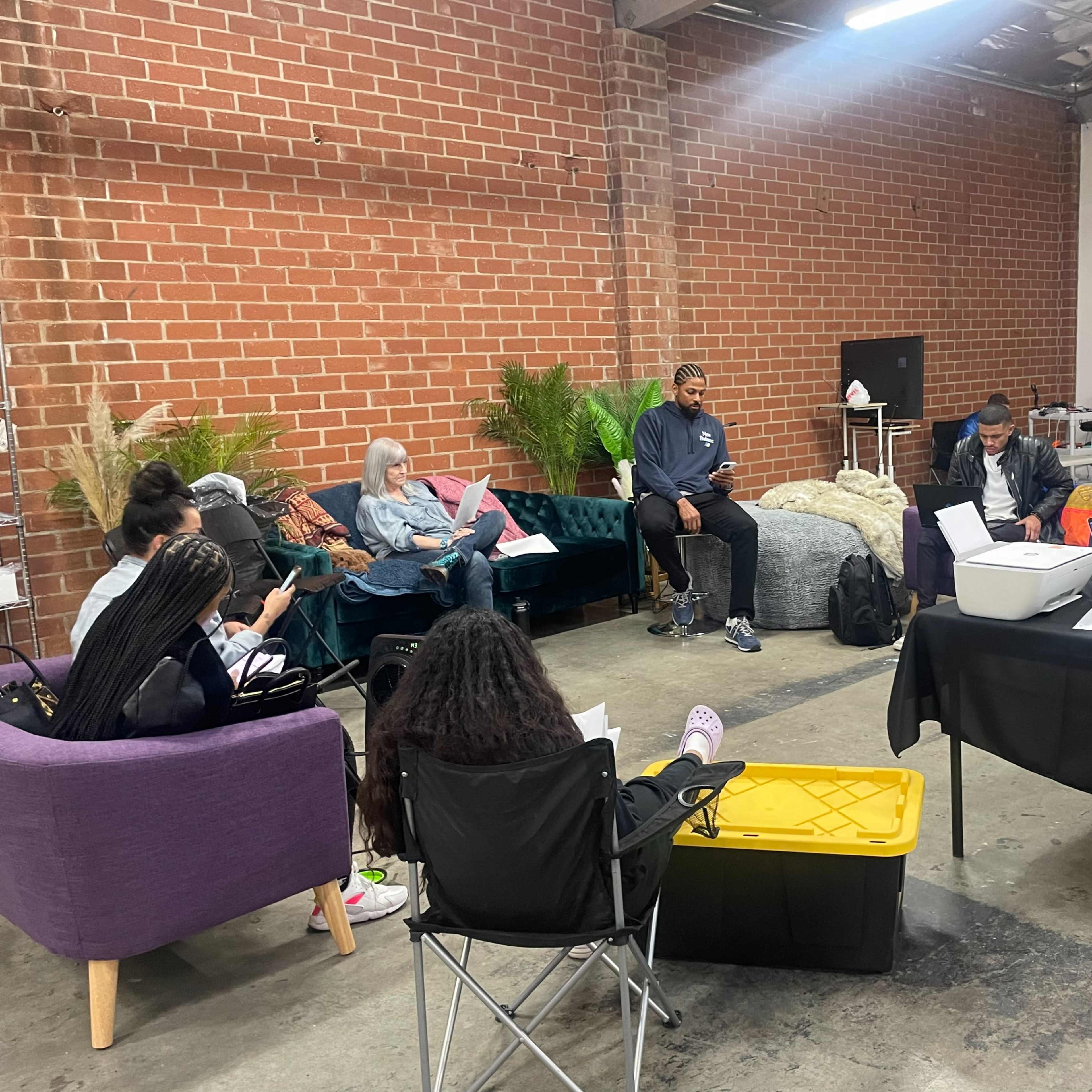 A group of individuals sits in a workshop setting, with some engaged in discussions and others using laptops, surrounded by modern furnishings in a brick-walled room.