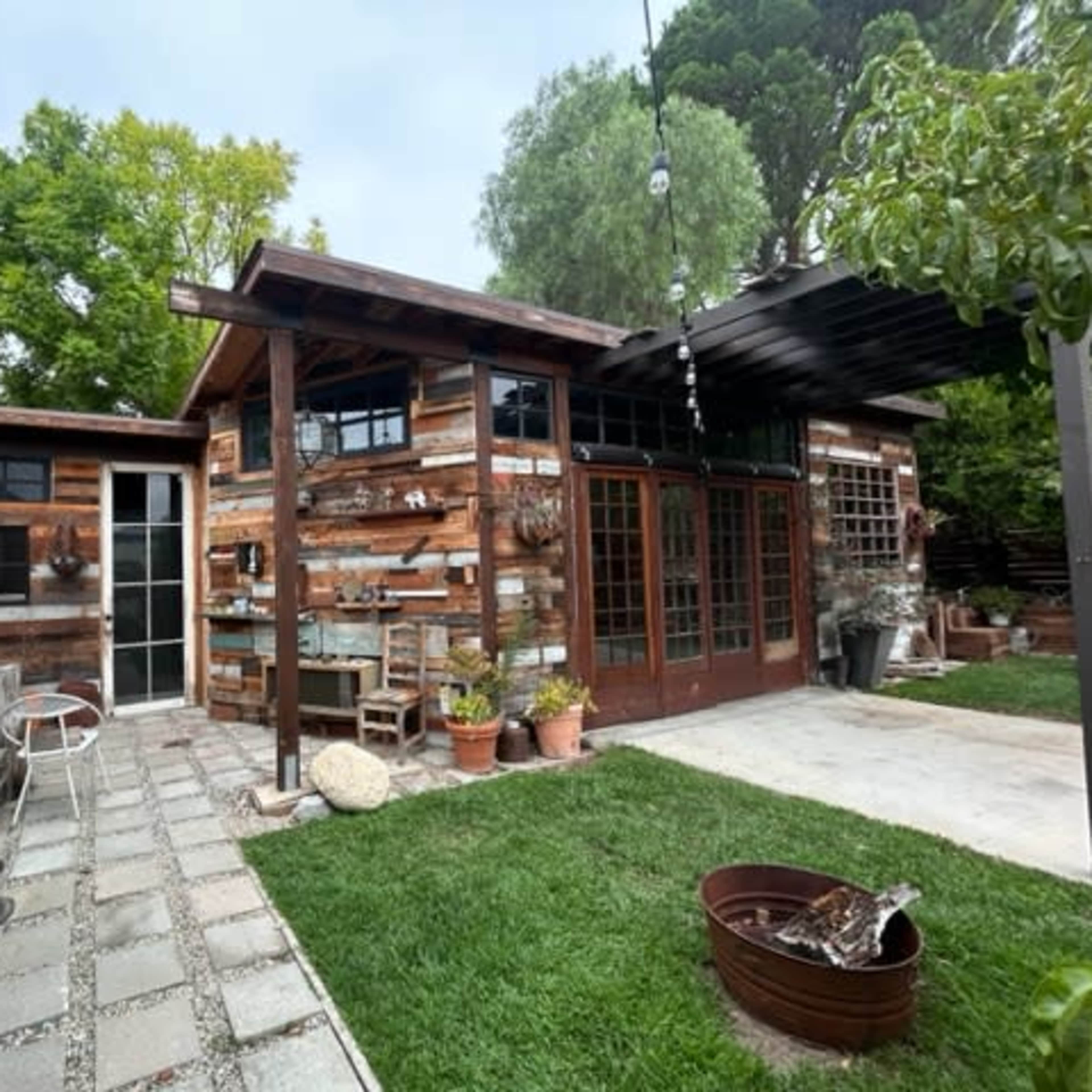 A rustic wooden structure situated next to a stone-paved patio and surrounded by green grass and potted plants.