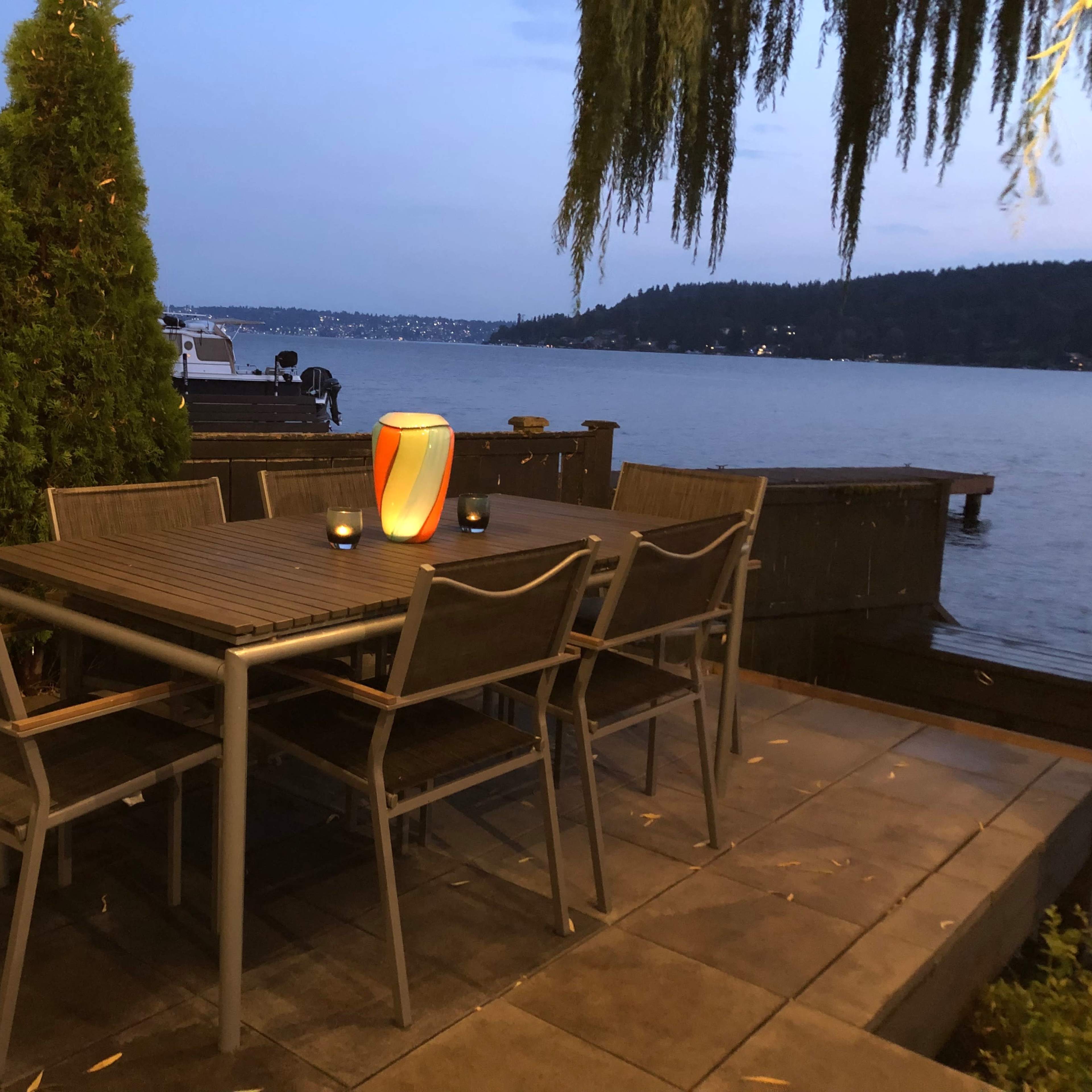 A wooden dining table with six chairs is set near the water's edge, illuminated by a lantern and overlooking a lake surrounded by trees and hills.