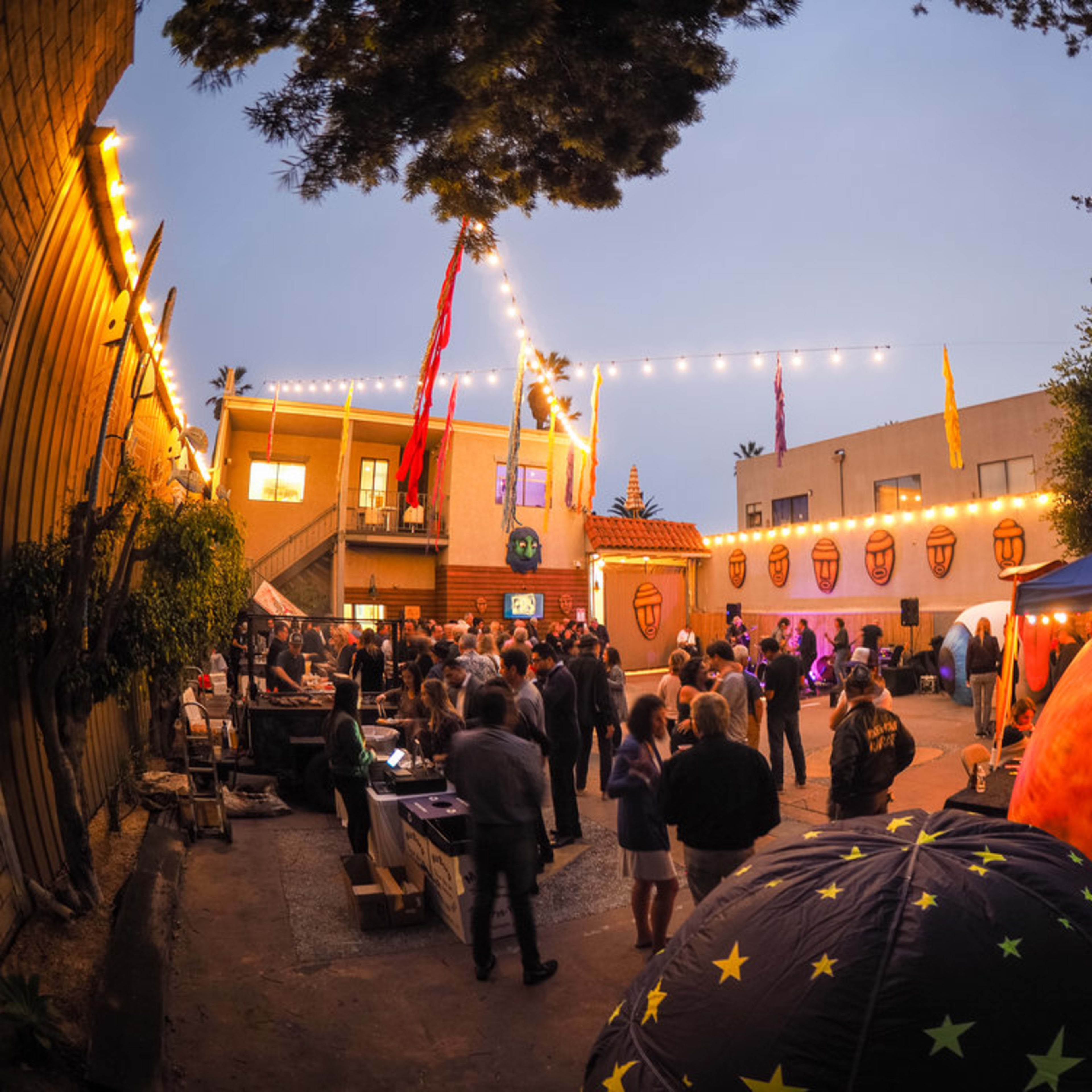 A crowd mingles in an outdoor space decorated with lights and banners during a social event in the evening.