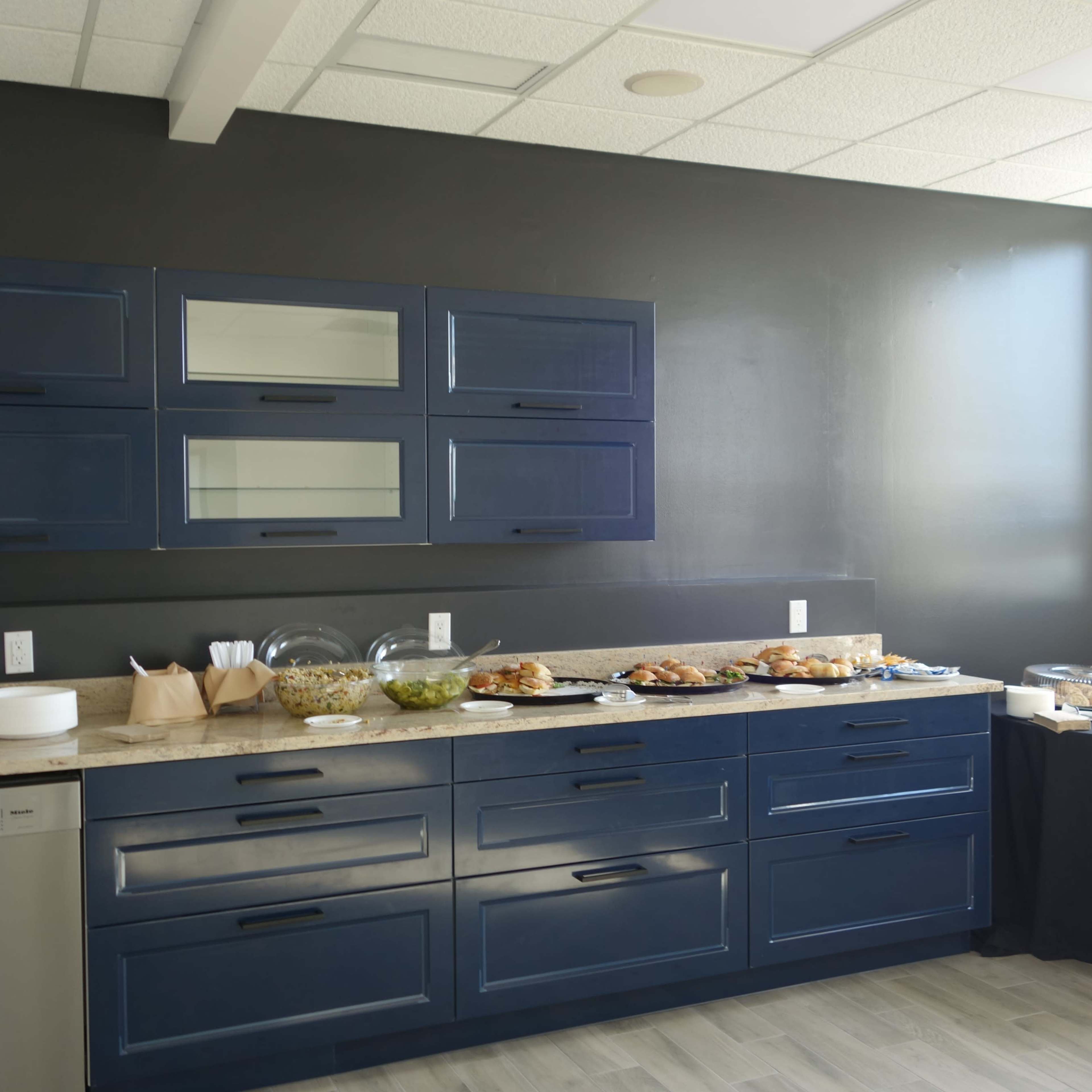 A modern kitchen space features navy blue cabinets, a stone countertop, and a variety of food items displayed on a table.