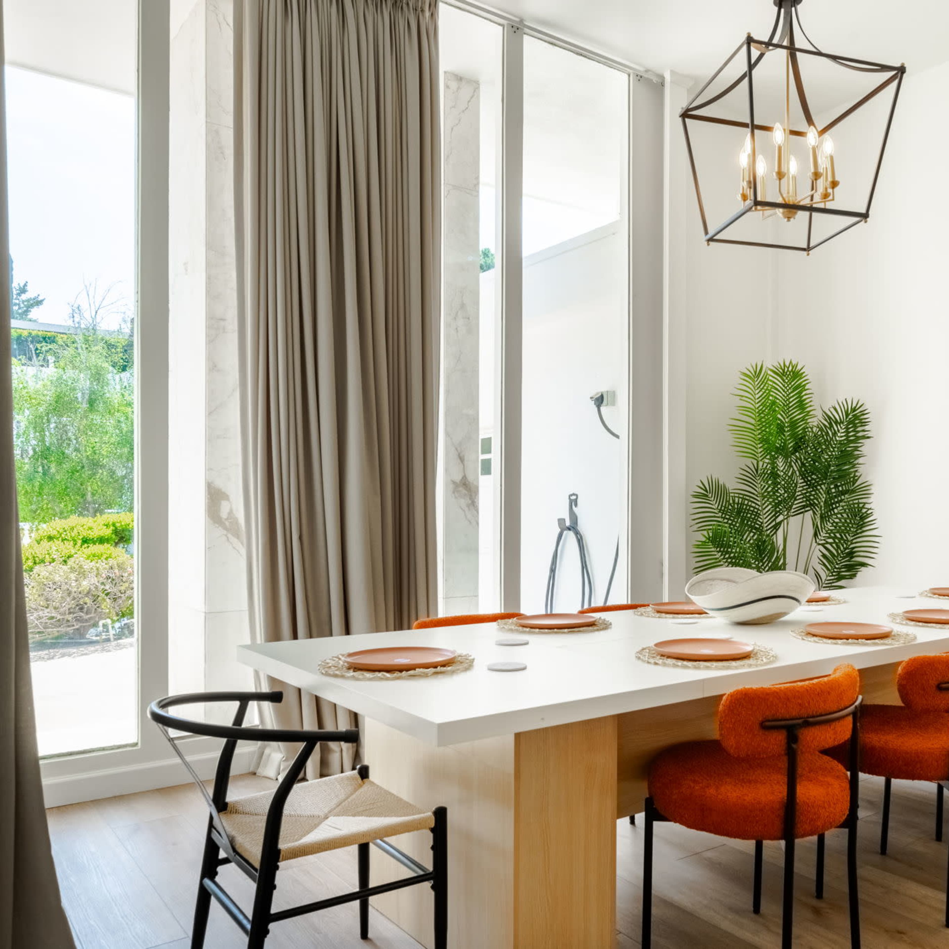 A modern dining area features a long table with orange chairs, warm wood flooring, and large windows allowing natural light.