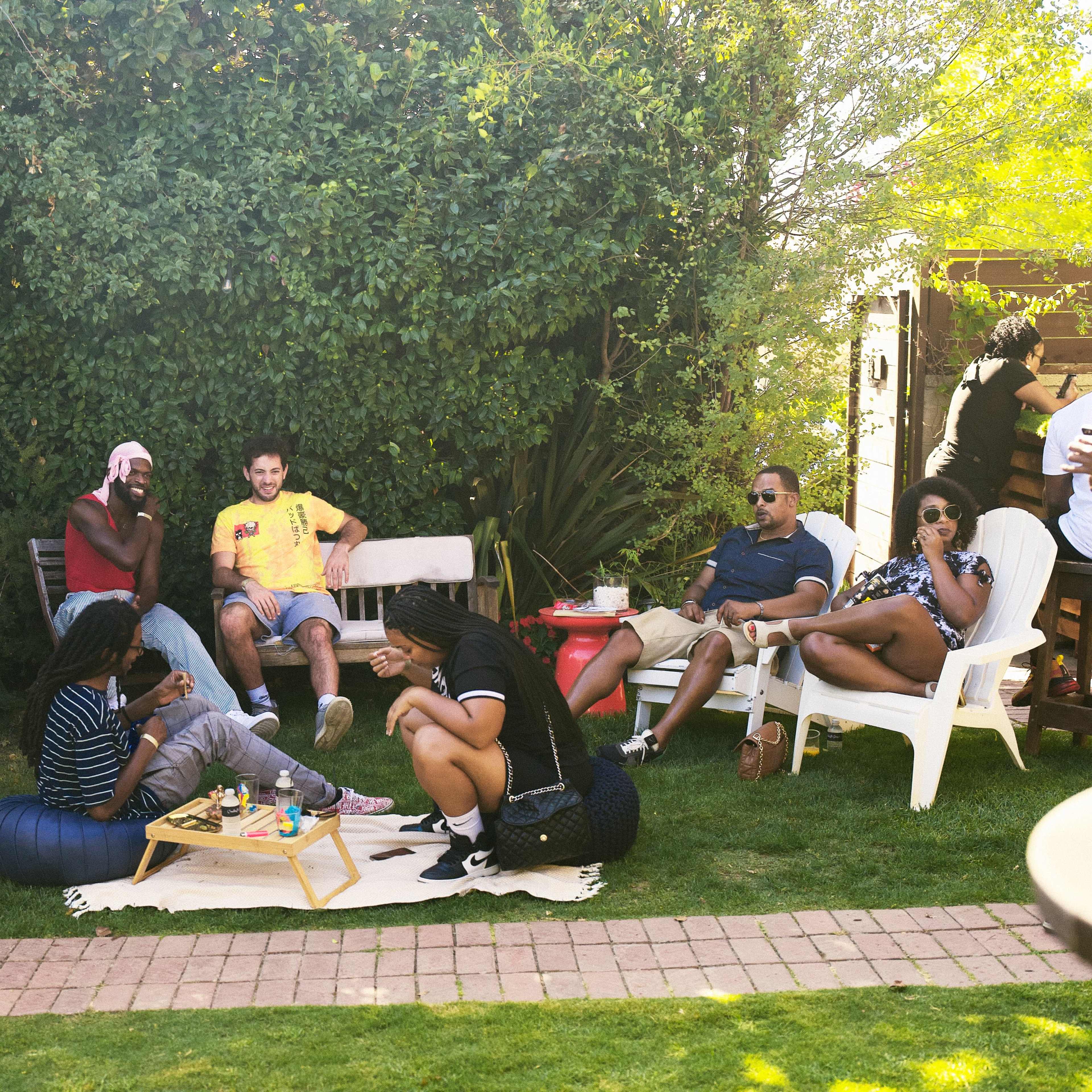 A group of people gathers in a backyard garden, sitting on various chairs and lounging on the grass, while sharing food and drinks.