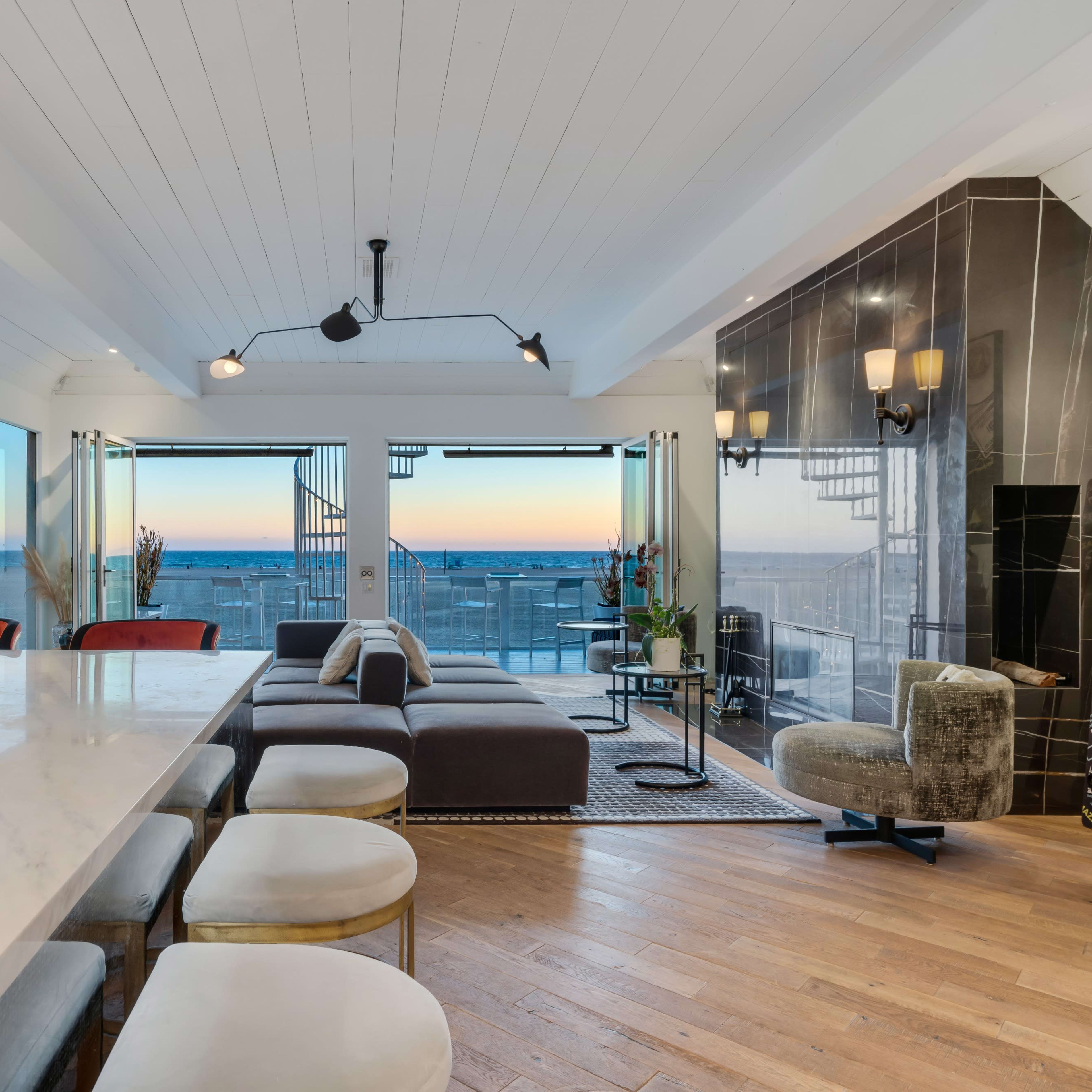 A modern living area with a large marble dining table, a sectional sofa, and expansive windows showcasing a sunset view.