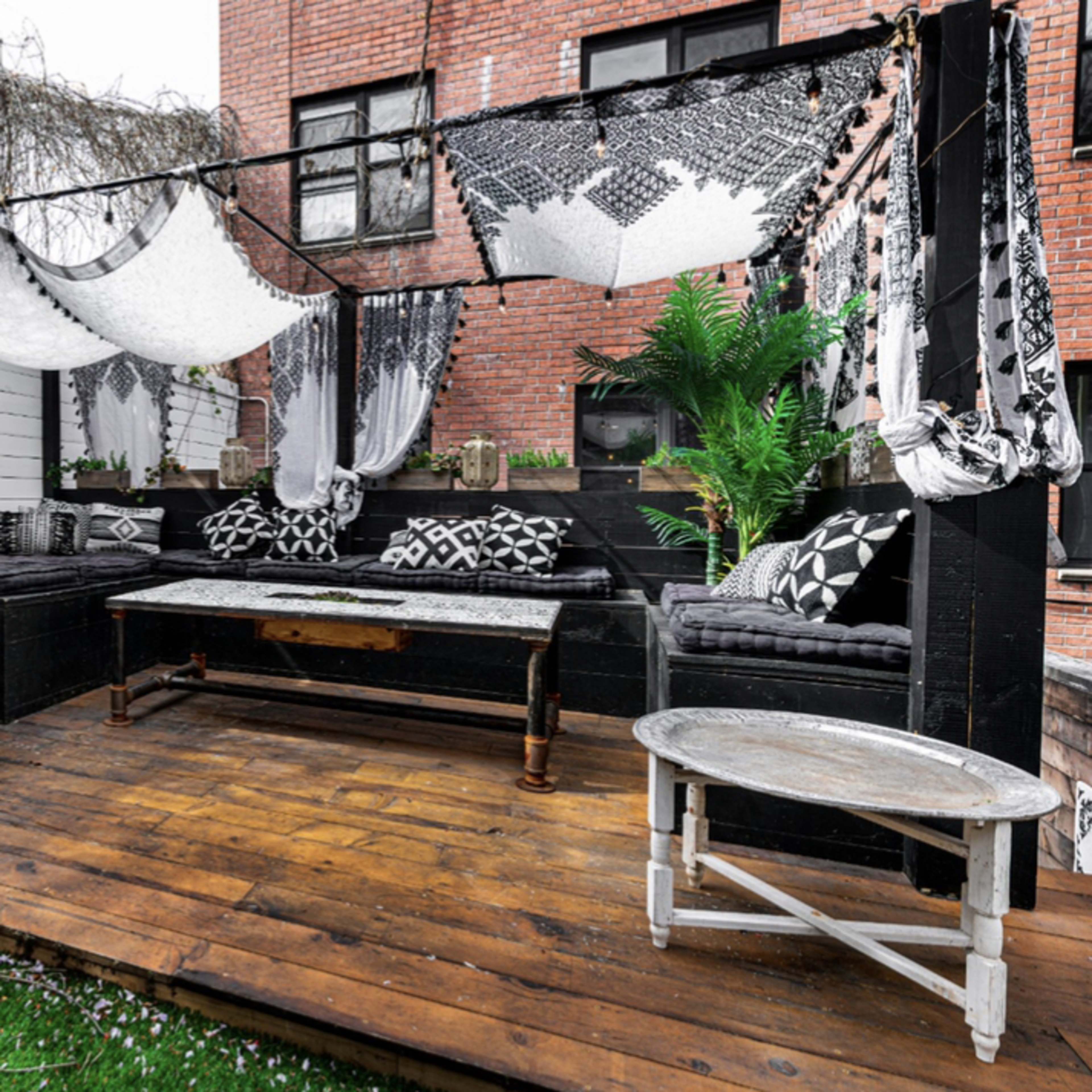 A patio features black and white seating, decorative textiles, potted plants, and wooden flooring.
