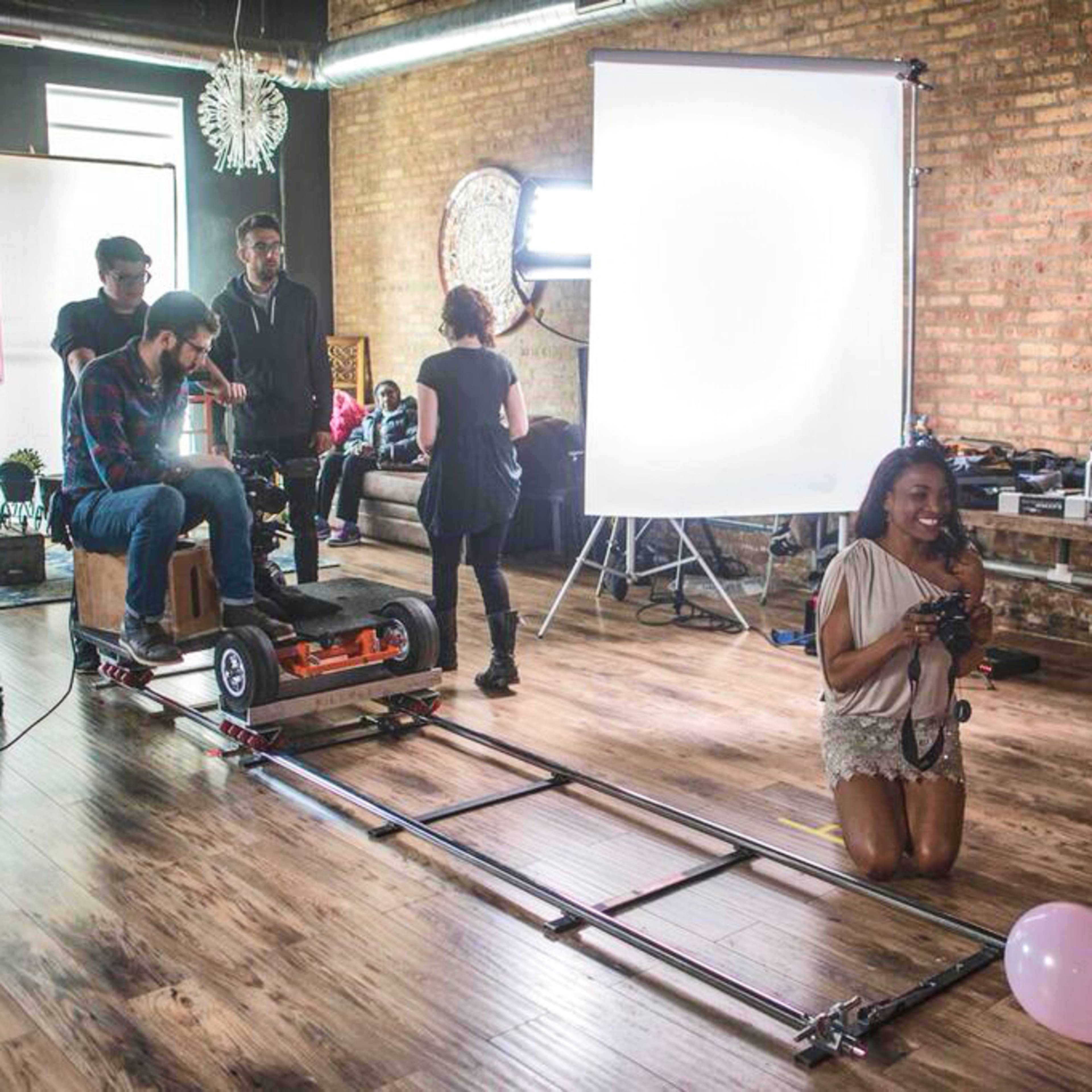 A film crew sets up a scene in a studio with a camera on a track, while actors prepare on a set featuring various equipment and props.