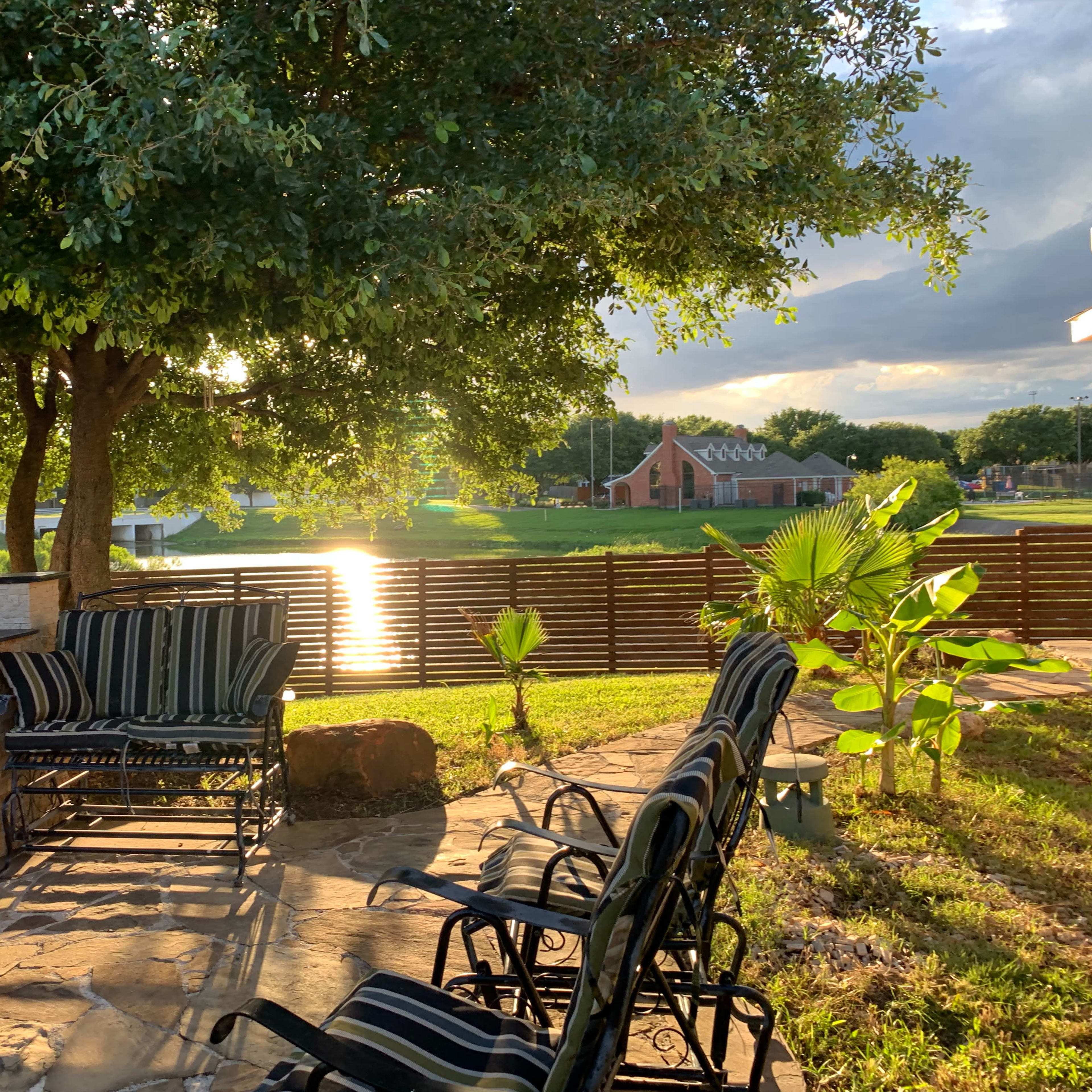A patio area with striped chairs and a stone barbecue overlooks a tranquil water surface reflecting the sunlight, surrounded by greenery.