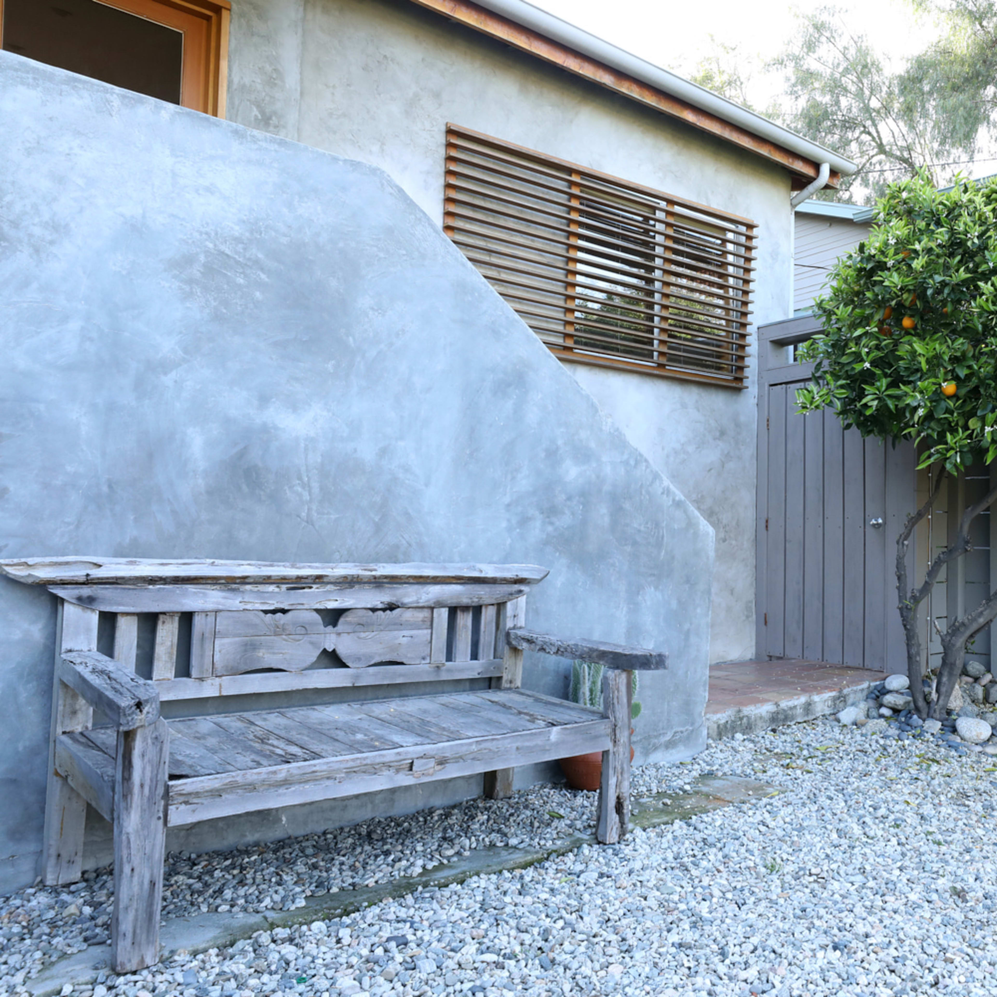 A weathered wooden bench sits on gravel next to a wall with a window, alongside a tree bearing oranges.