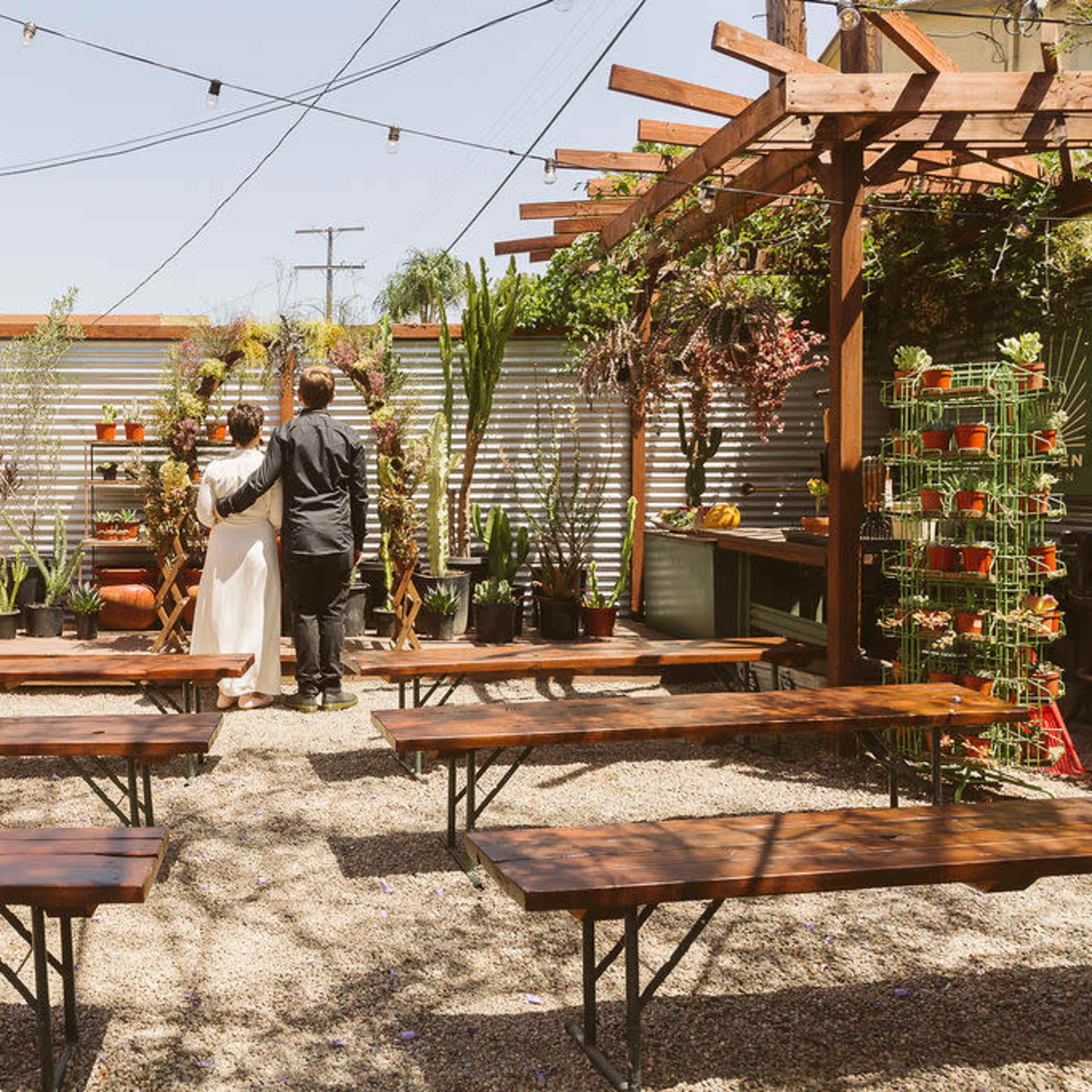 A couple stands together in a garden setting with wooden benches and various potted plants arranged along a metal backdrop.