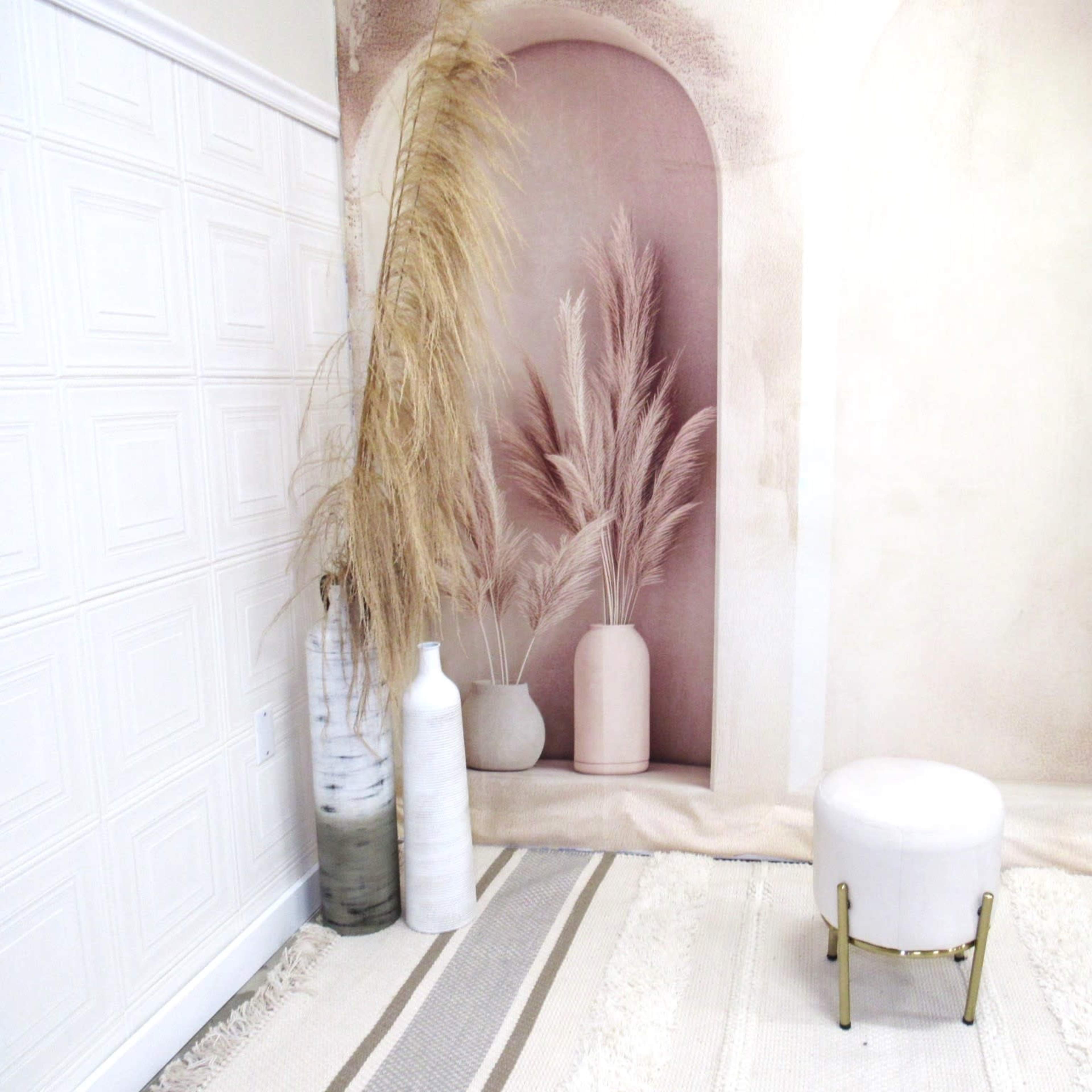 A styled interior scene featuring tall dried plants in vases, a small round stool, and a textured rug against a backdrop with arched shapes.