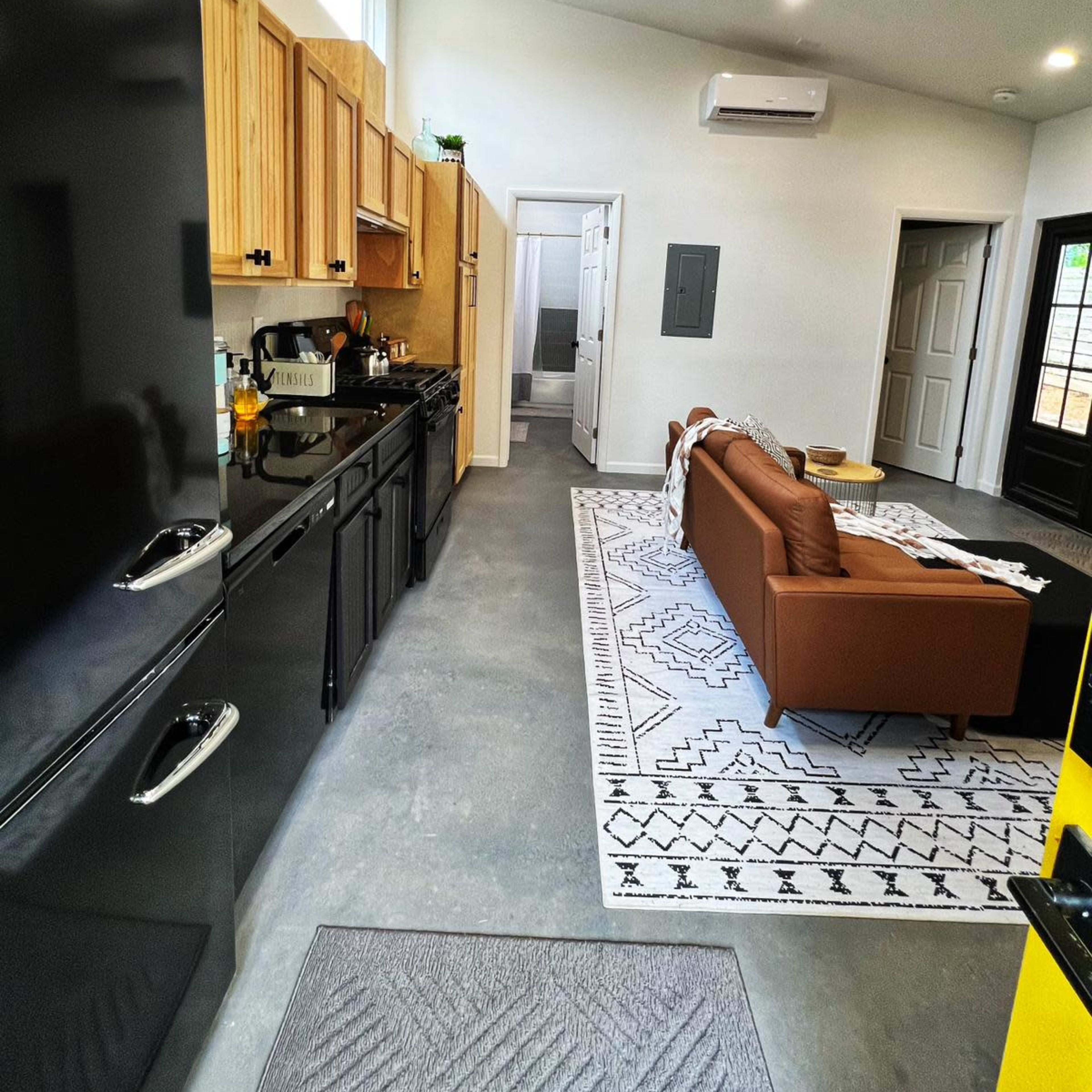 A modern kitchen and living area, featuring a black refrigerator, wooden cabinets, and a brown sofa on a patterned rug.
