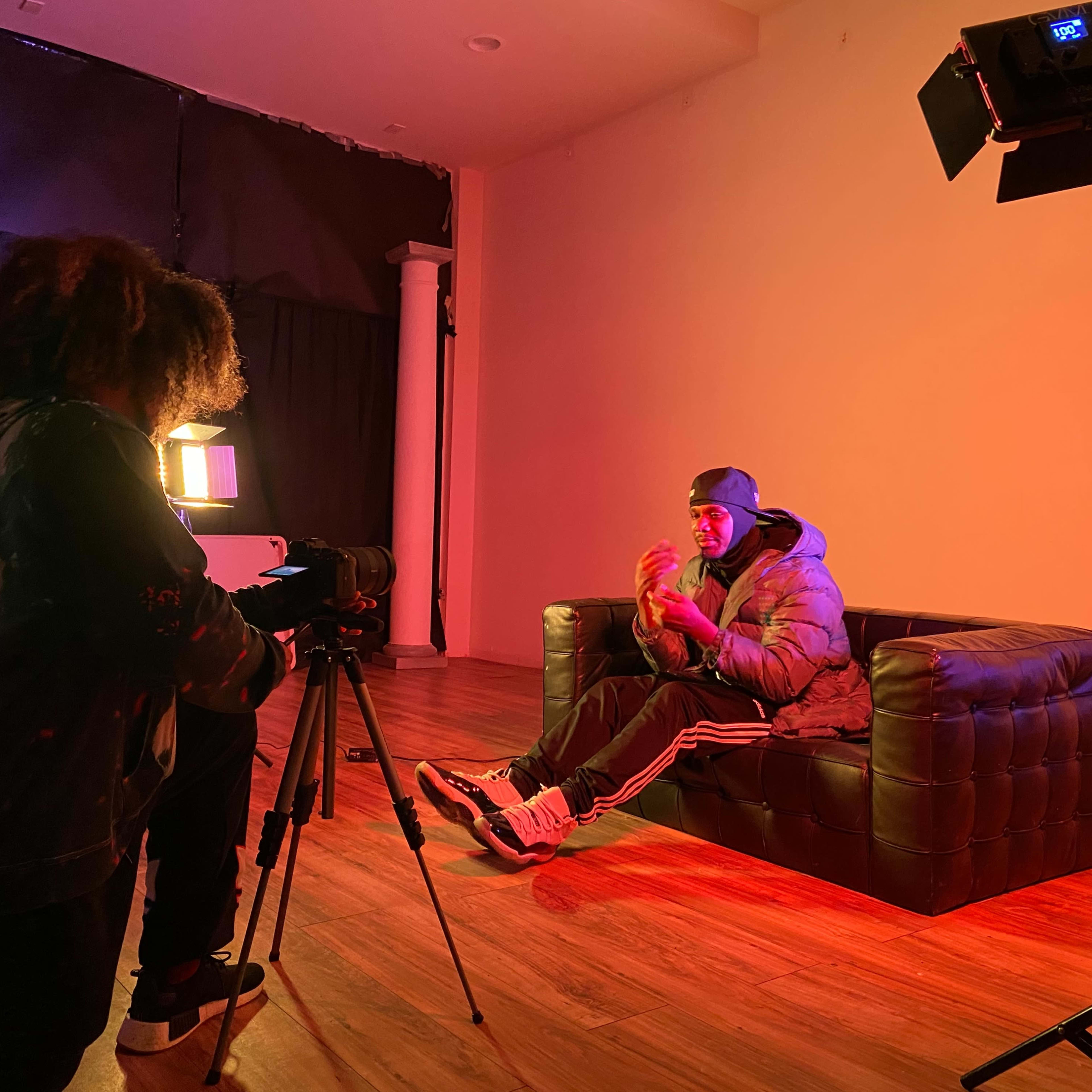 A person sits on a dark leather couch in a dimly lit room while another person operates a camera on a tripod, capturing the scene.