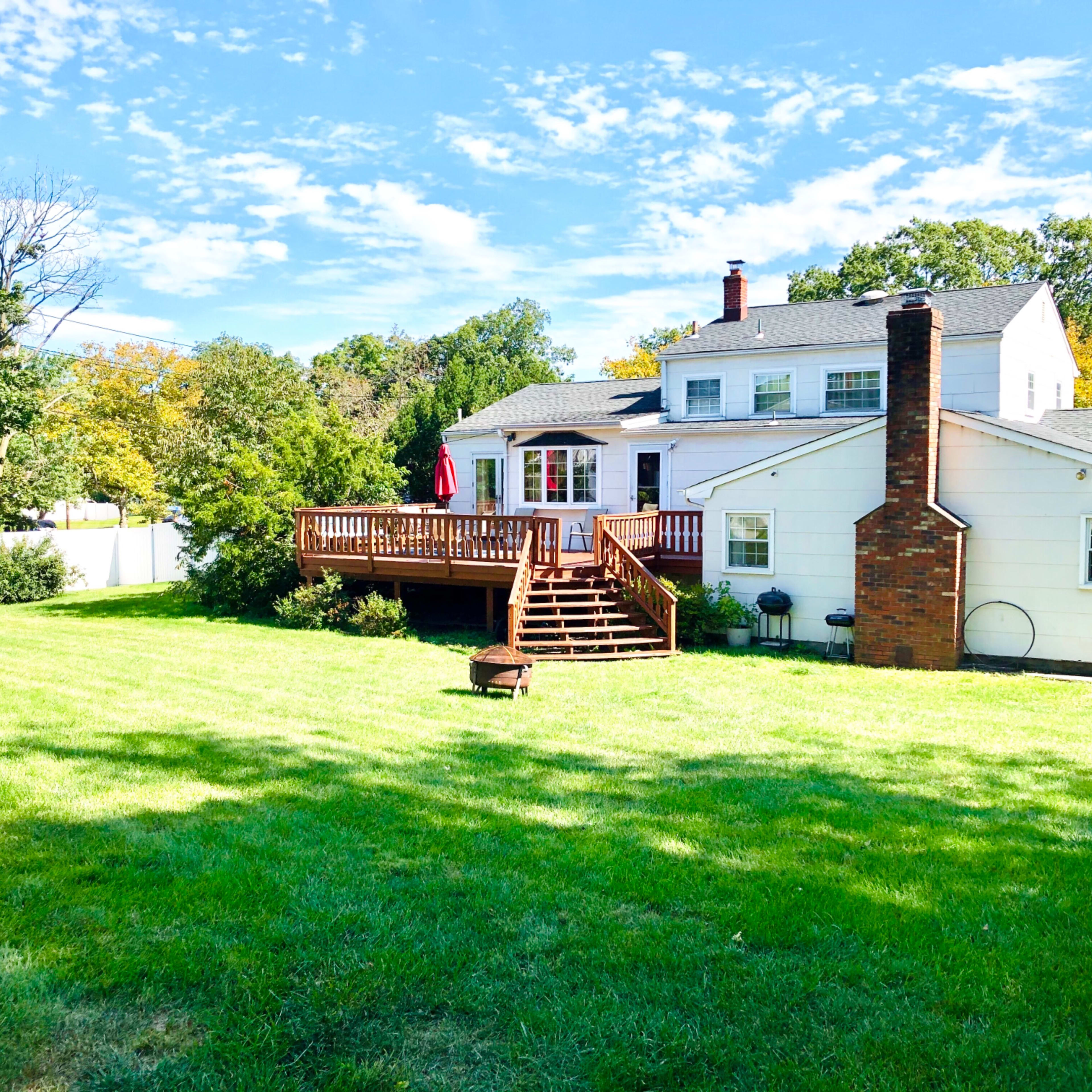 A spacious backyard with a wooden deck attached to a white house, surrounded by green grass and trees under a partly cloudy sky.