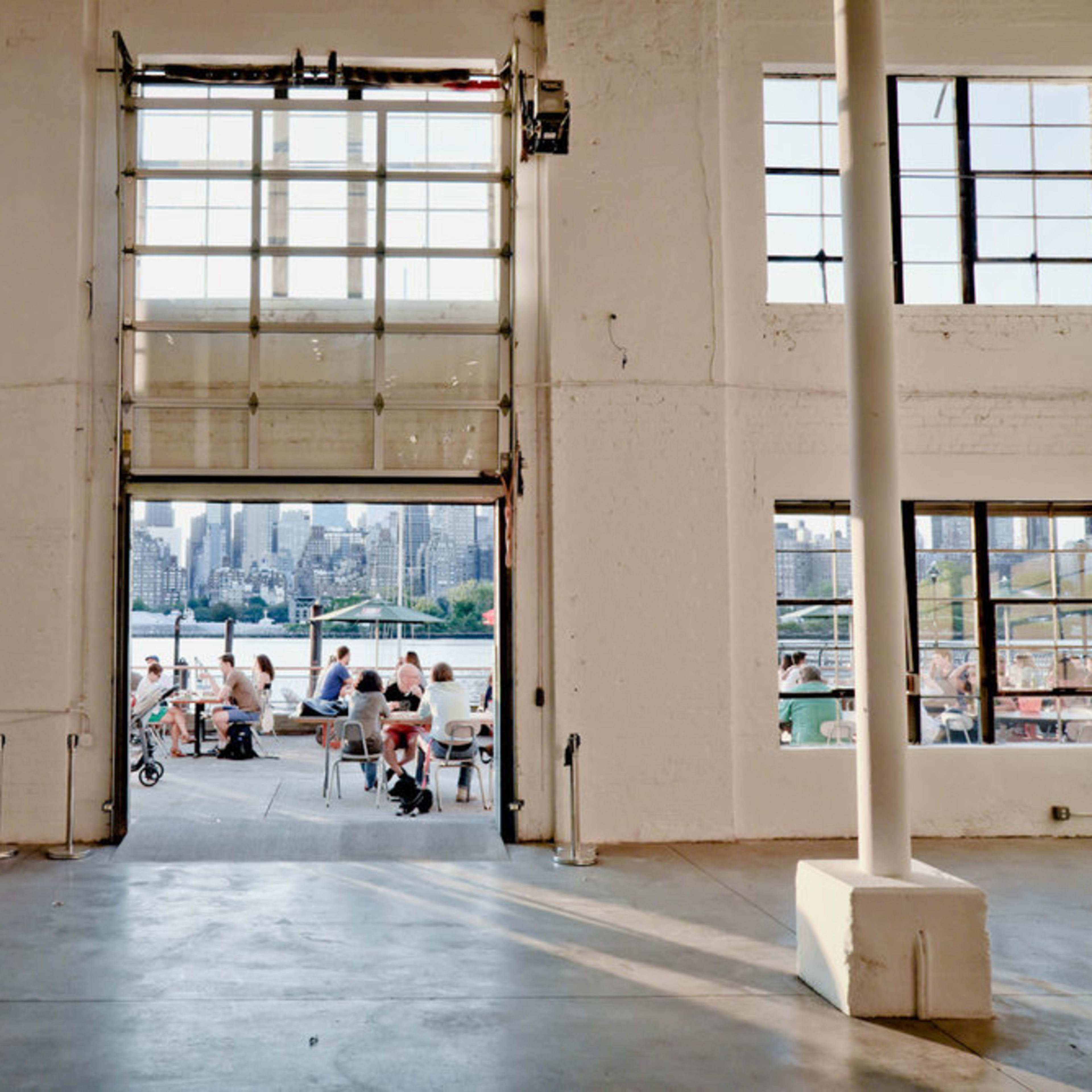 An open industrial-style entrance with large glass doors, revealing a view of a bustling outdoor area filled with people and the city skyline in the background.