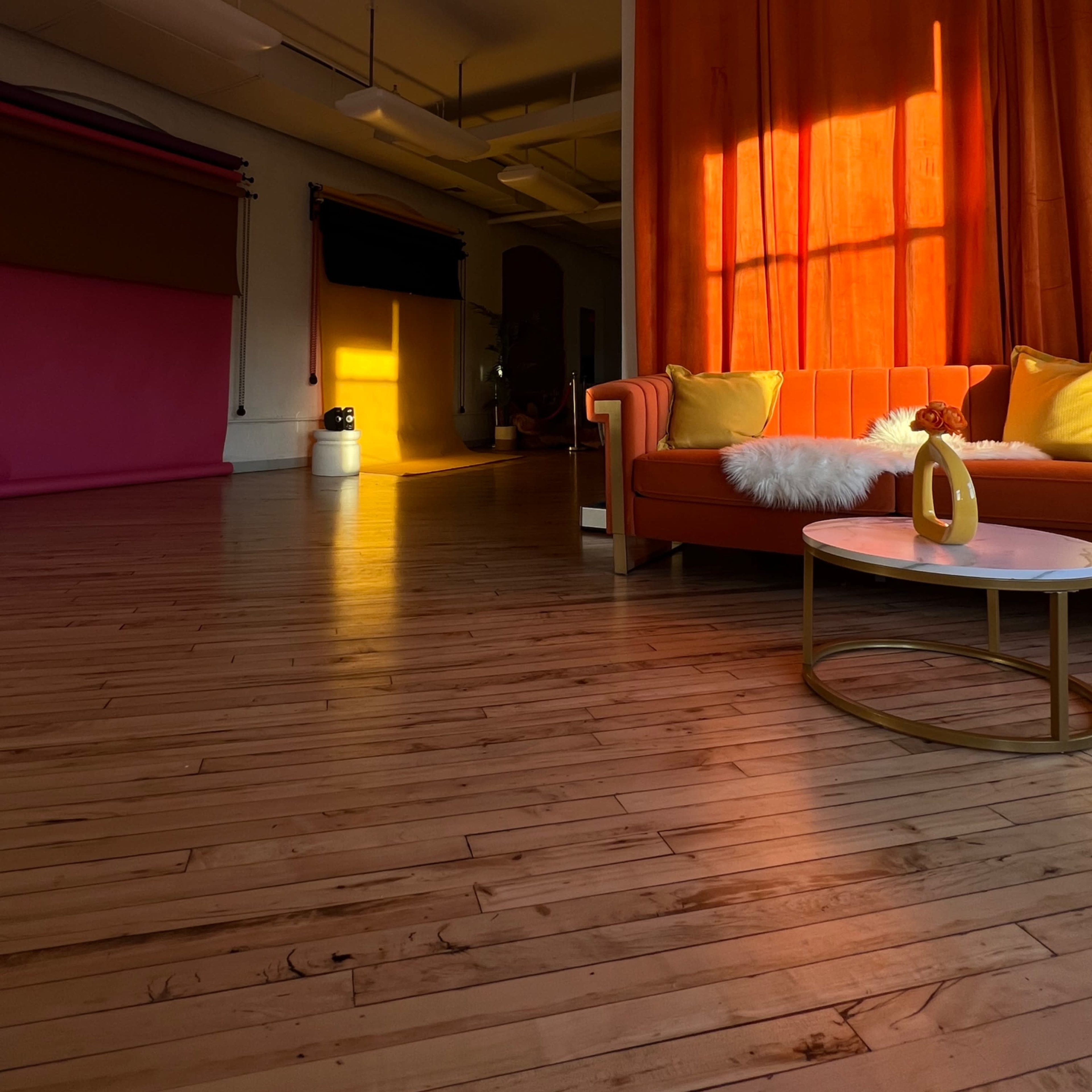 A well-lit interior features an orange couch with decorative pillows, a round white table, and large orange curtains, set against a wooden floor.