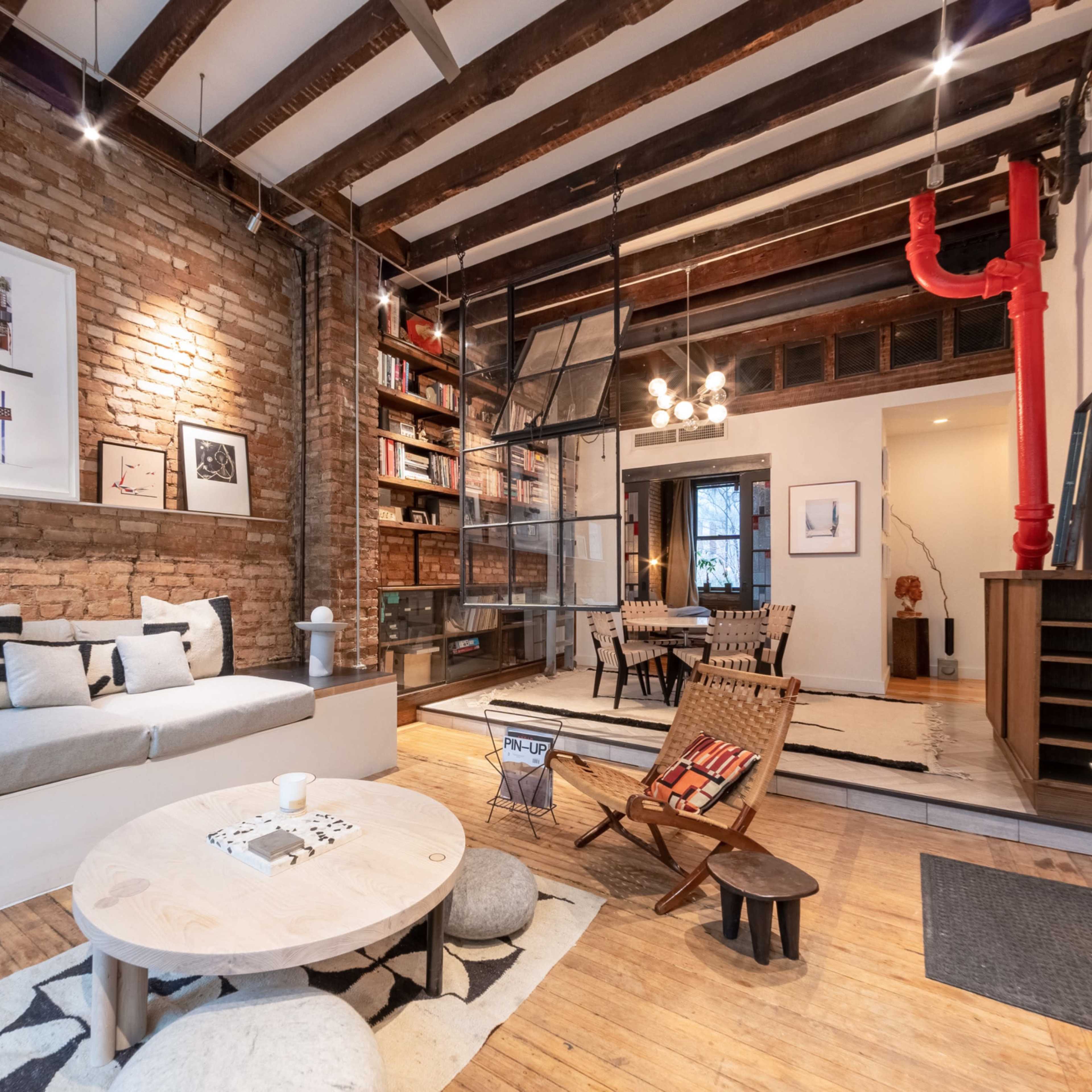 A stylish living room with exposed brick walls, wooden beams, and a mix of modern furniture, including a long couch, a round coffee table, and a wooden chair.