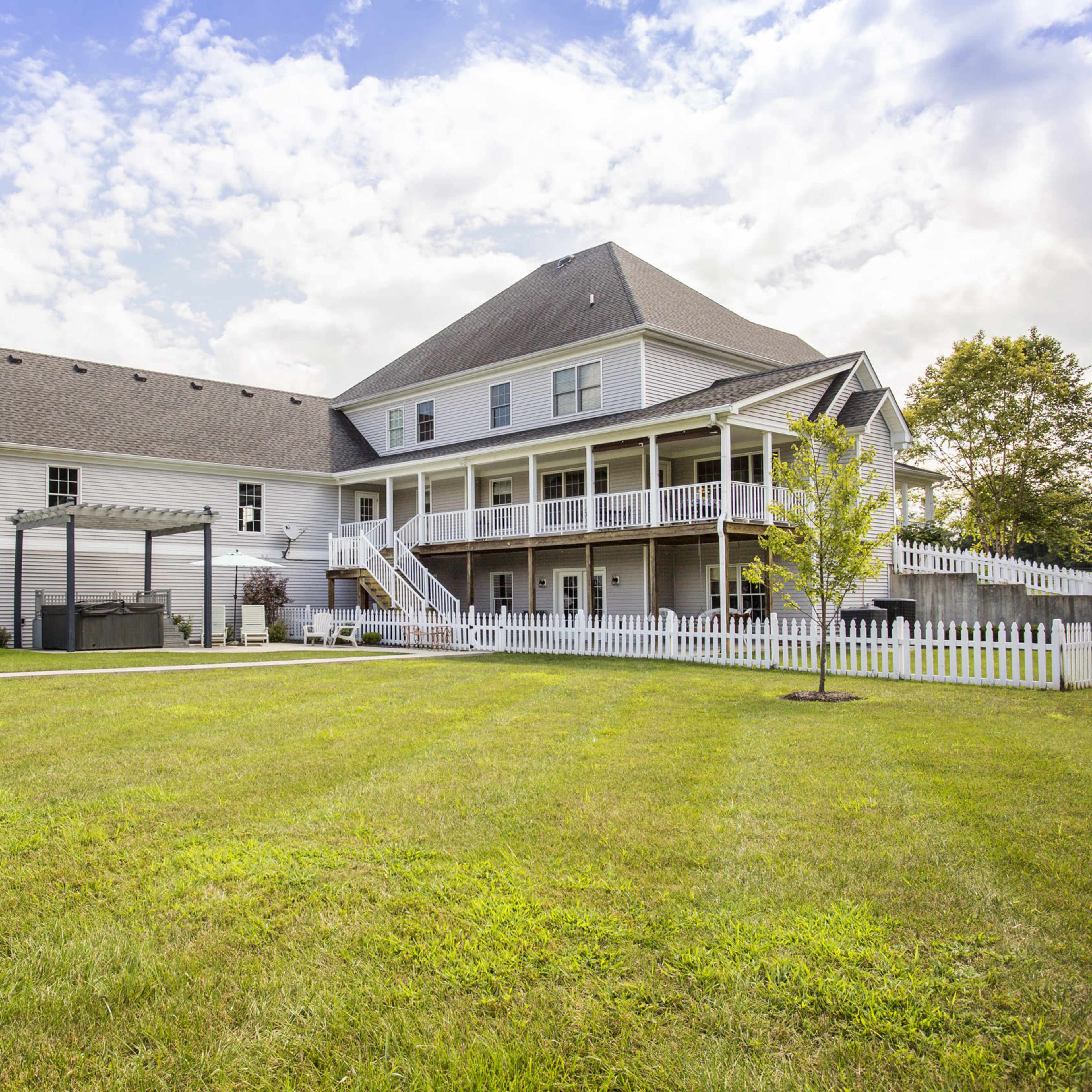 A large two-story house with a peaked roof and a wraparound porch is set on a grassy lawn surrounded by a white picket fence.