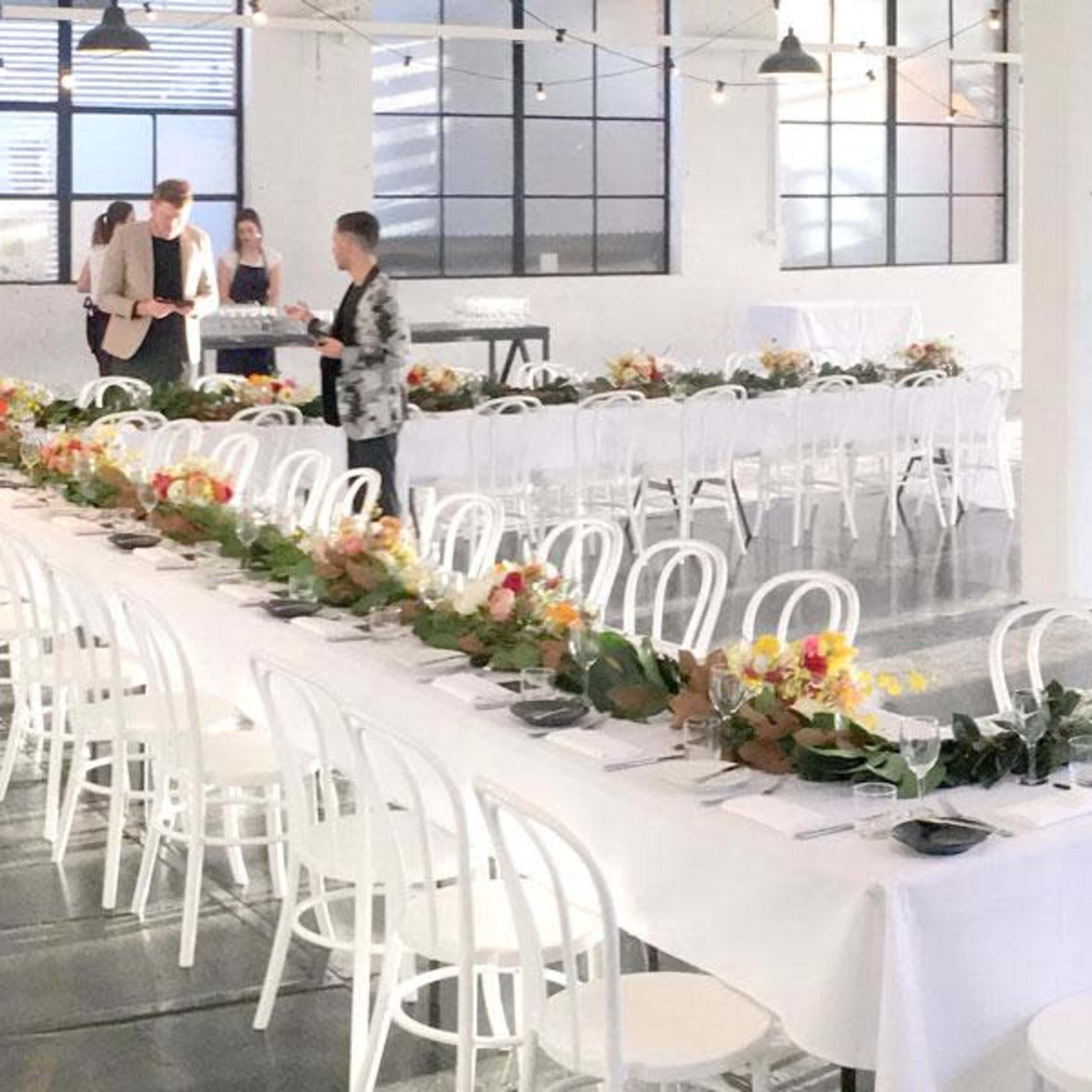 A long banquet table is set in a bright, open space, decorated with floral arrangements and surrounded by white chairs.