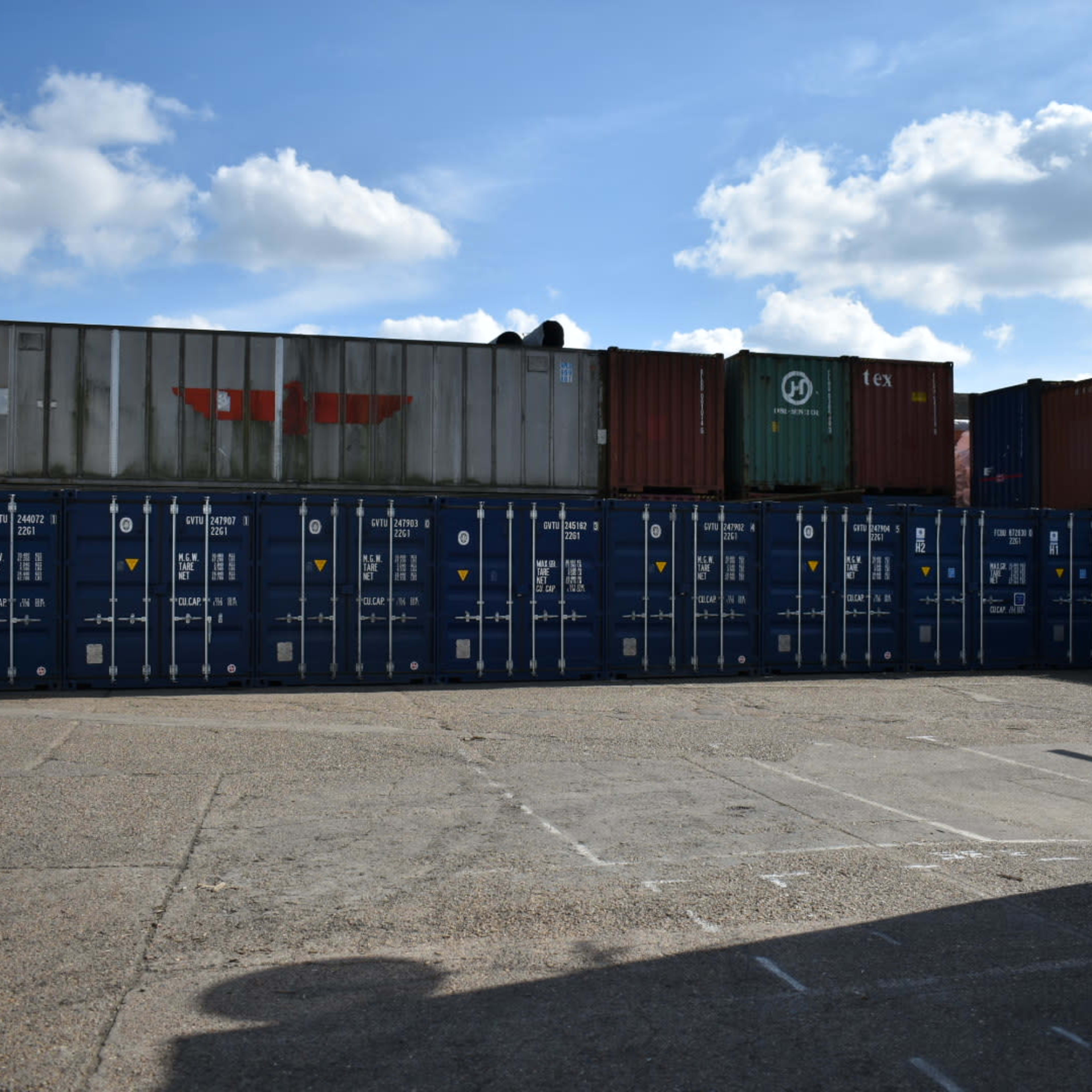 A row of blue shipping containers stacked against a cloudy sky in an industrial area.