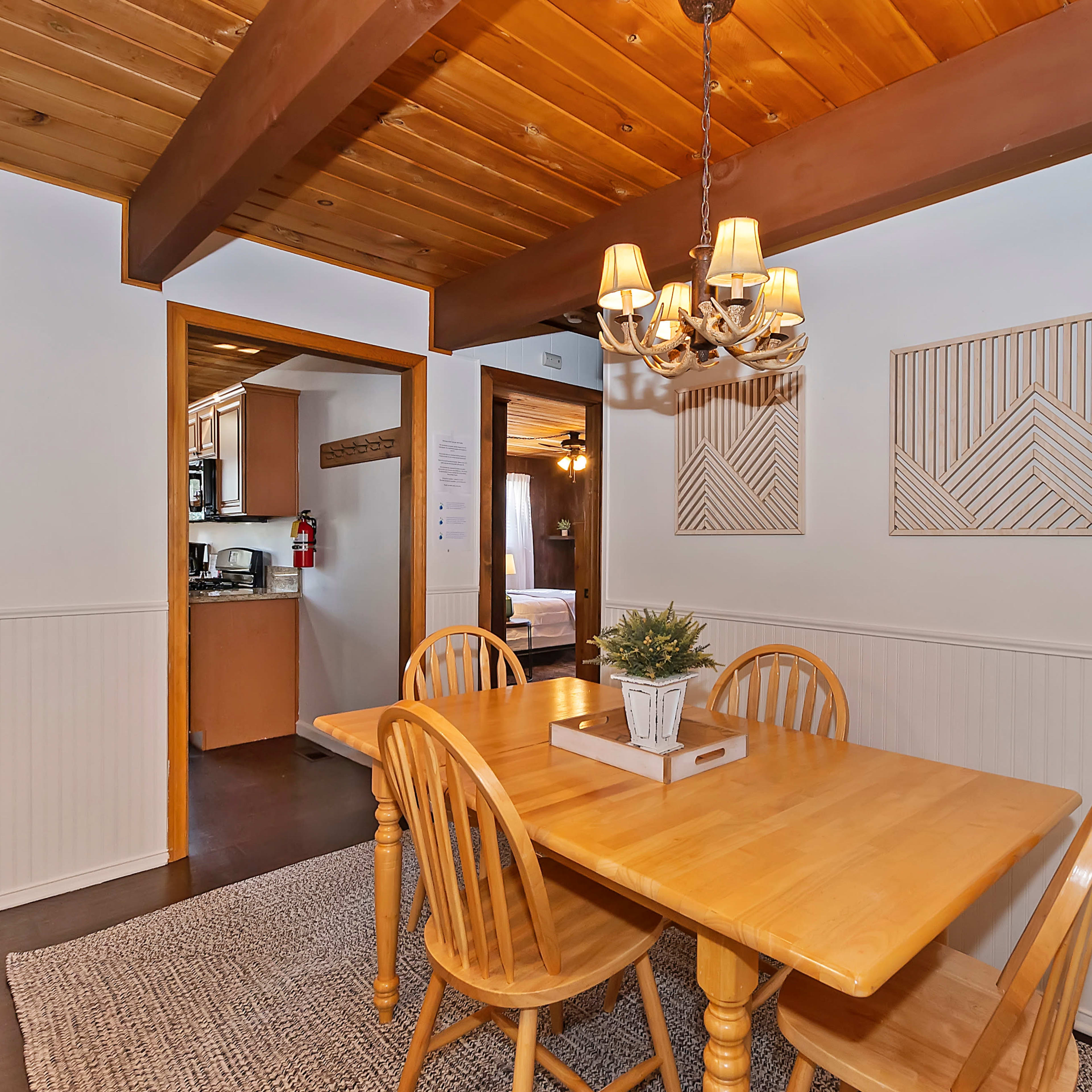 A wooden dining table with four chairs is positioned in a well-lit room featuring white walls and wooden beams overhead.