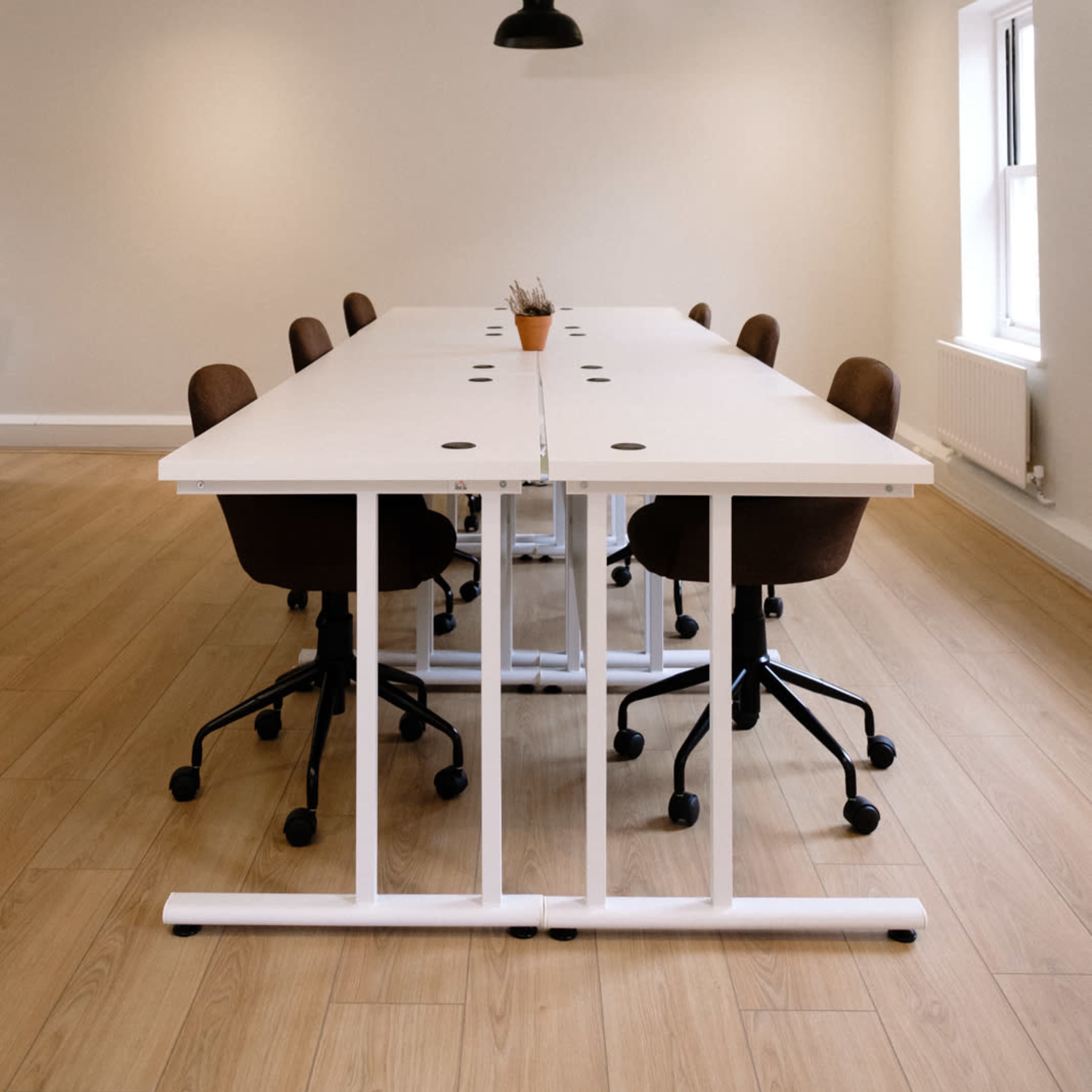 A modern meeting room features a long white table surrounded by brown office chairs, illuminated by black pendant lights.