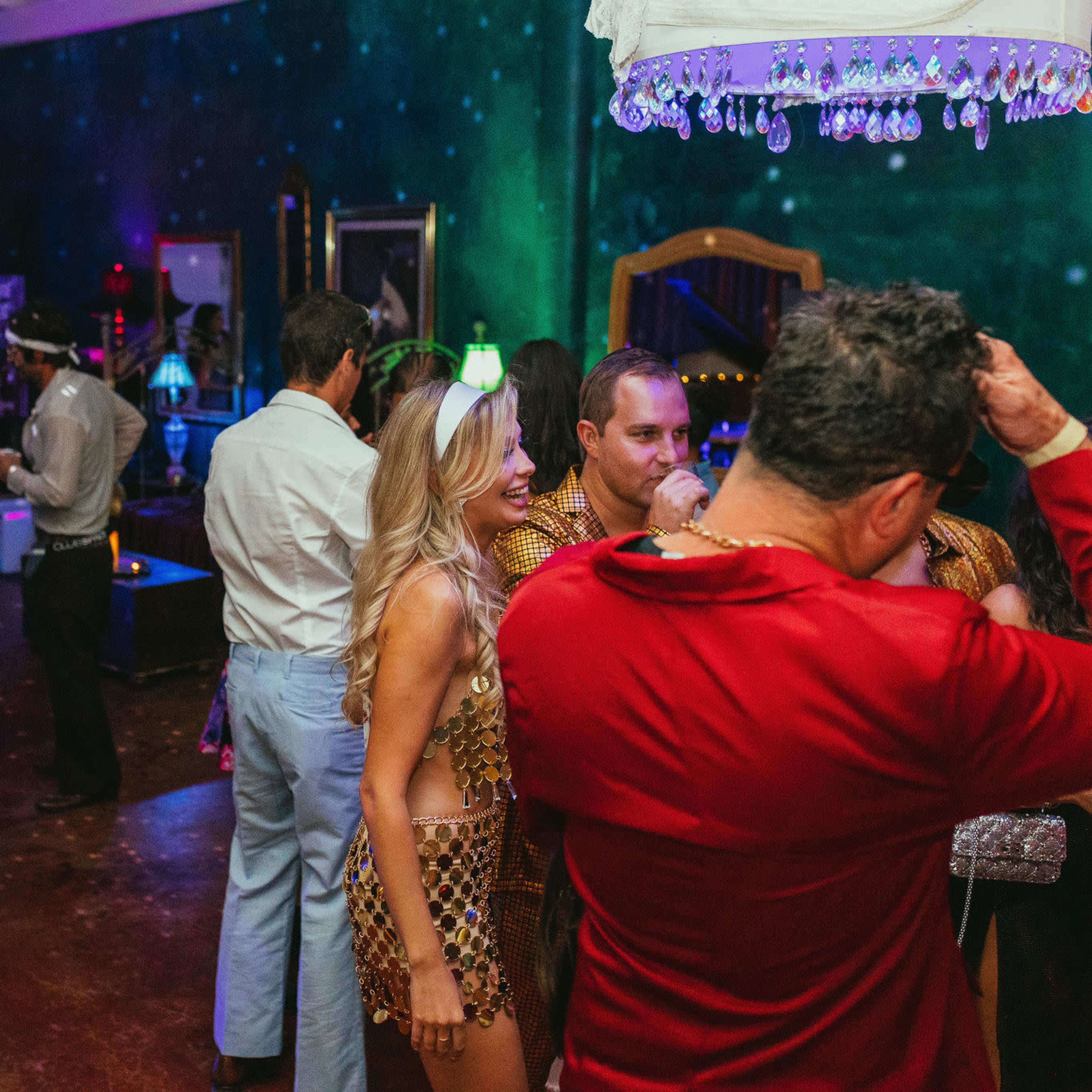 A vibrant party scene featuring guests dressed in various outfits socializing under disco lights with a disco ball overhead.