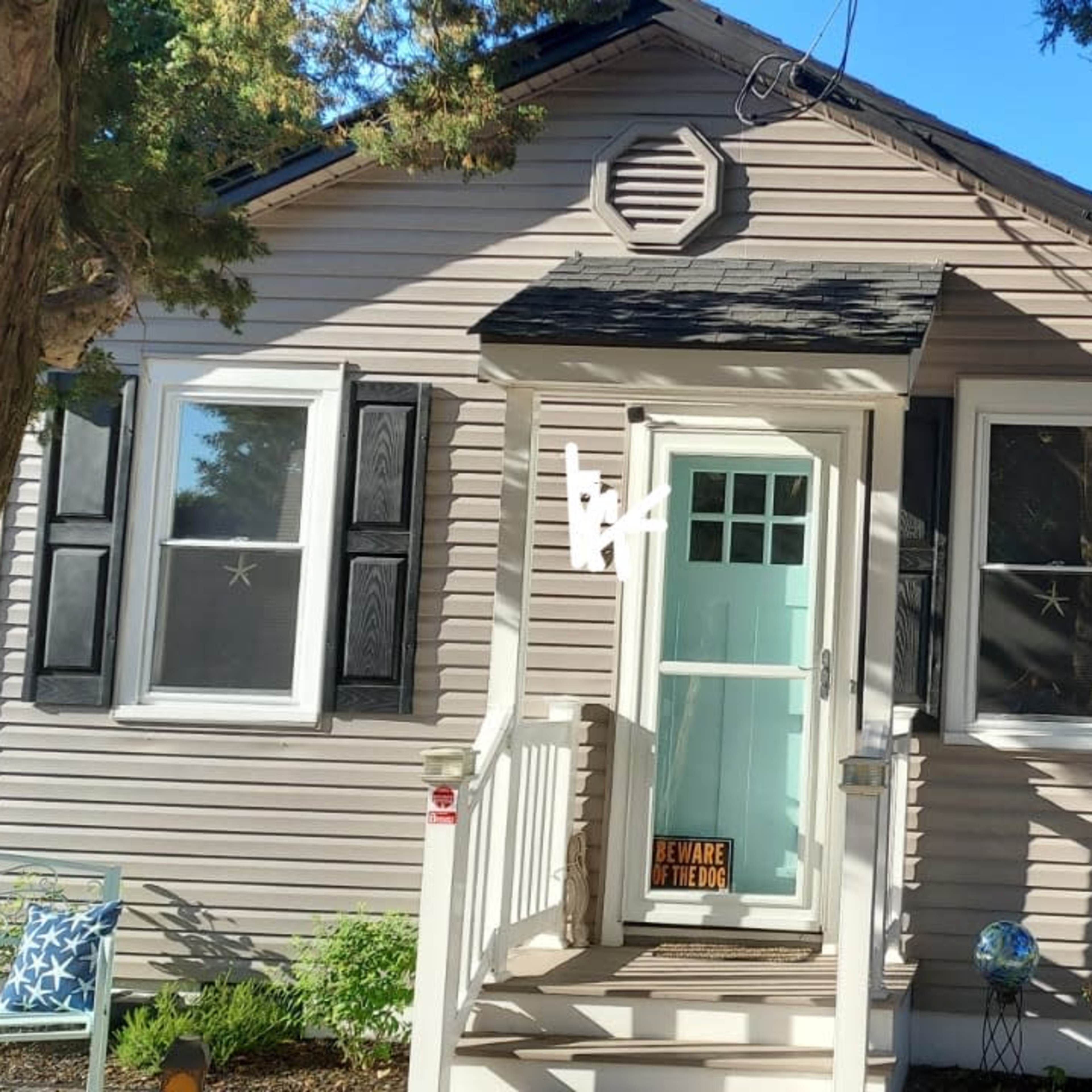 A small, single-story house with a light-colored exterior and black shutters is surrounded by trees and features a front porch with a welcoming sign.
