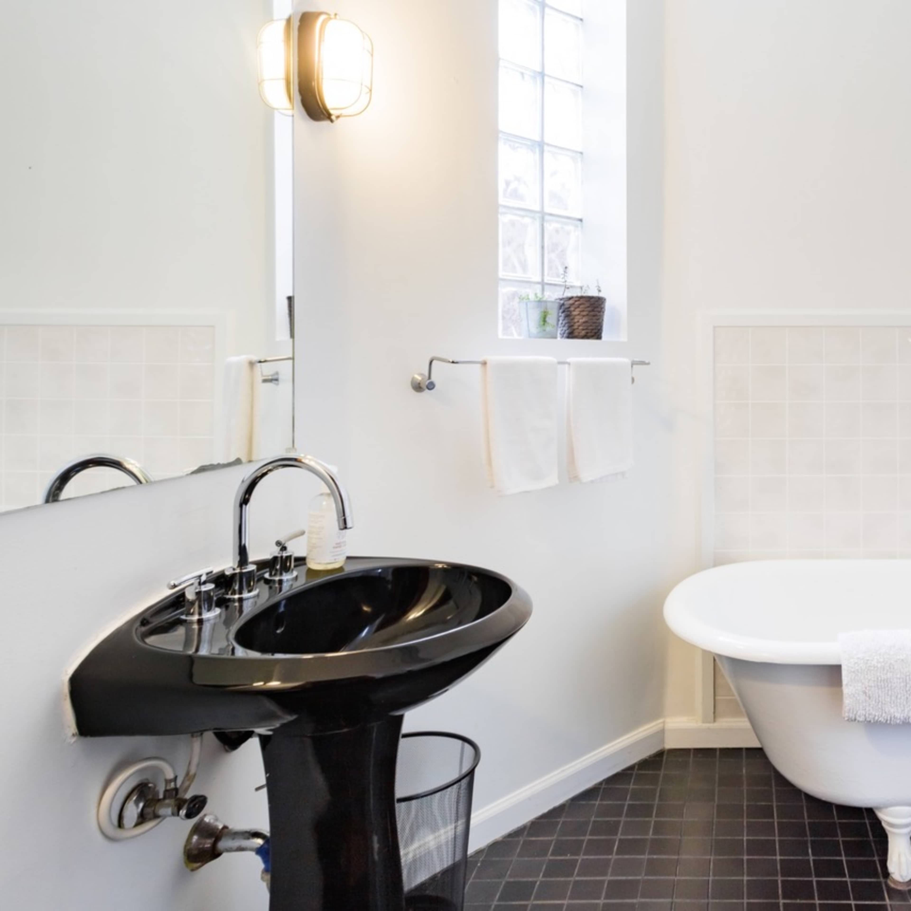 A modern bathroom featuring a black pedestal sink, a white freestanding bathtub, and a black toilet, with light-colored walls and tiled flooring.