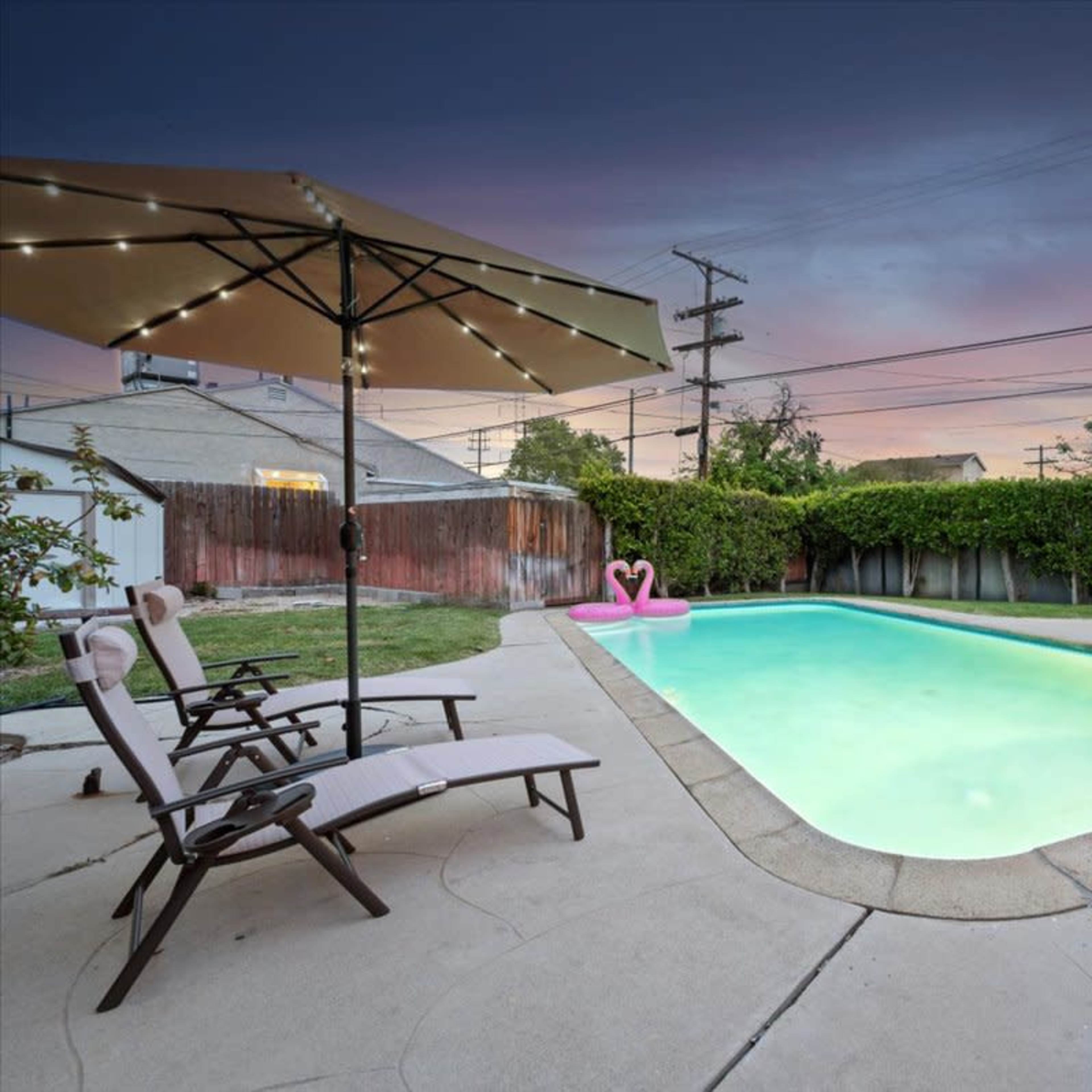 A backyard swimming pool surrounded by lounge chairs and umbrellas, with a pink float in the water and a twilight sky in the background.