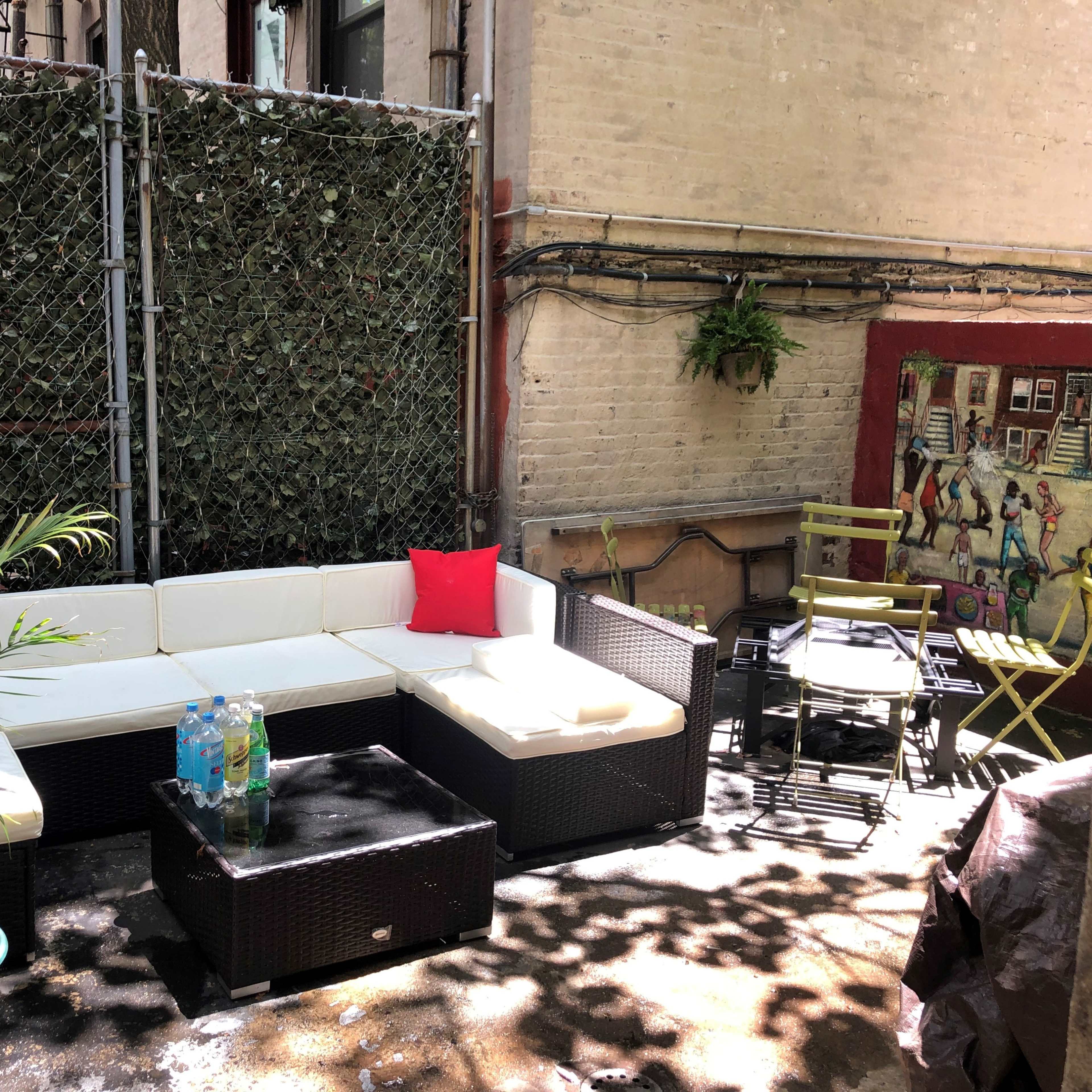 An outdoor seating area with a white sectional sofa, a glass-top coffee table, and a small dining table, surrounded by a chain-link fence and green foliage.