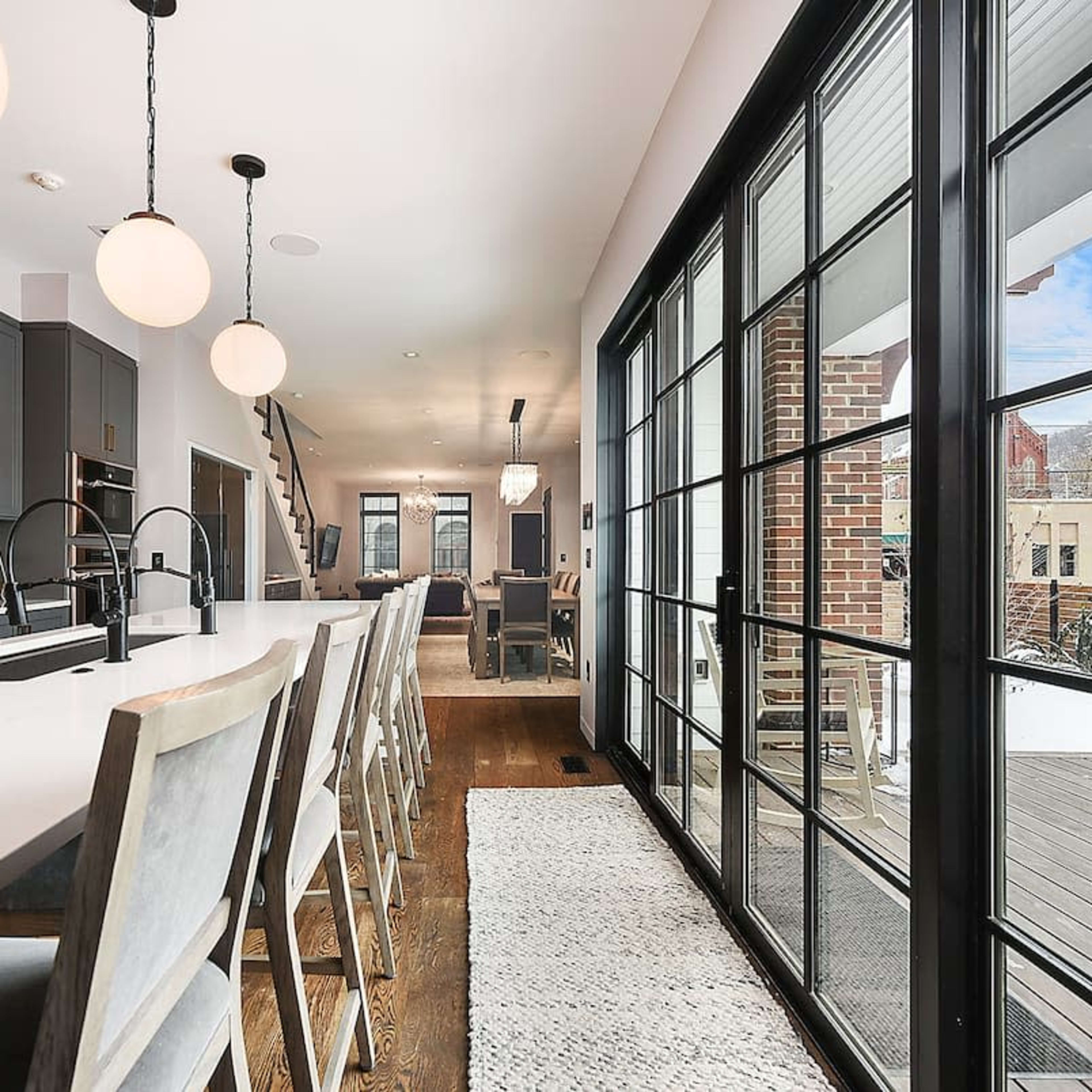 A modern kitchen with a long white countertop, gray cabinetry, and large windows leading to an outdoor space.