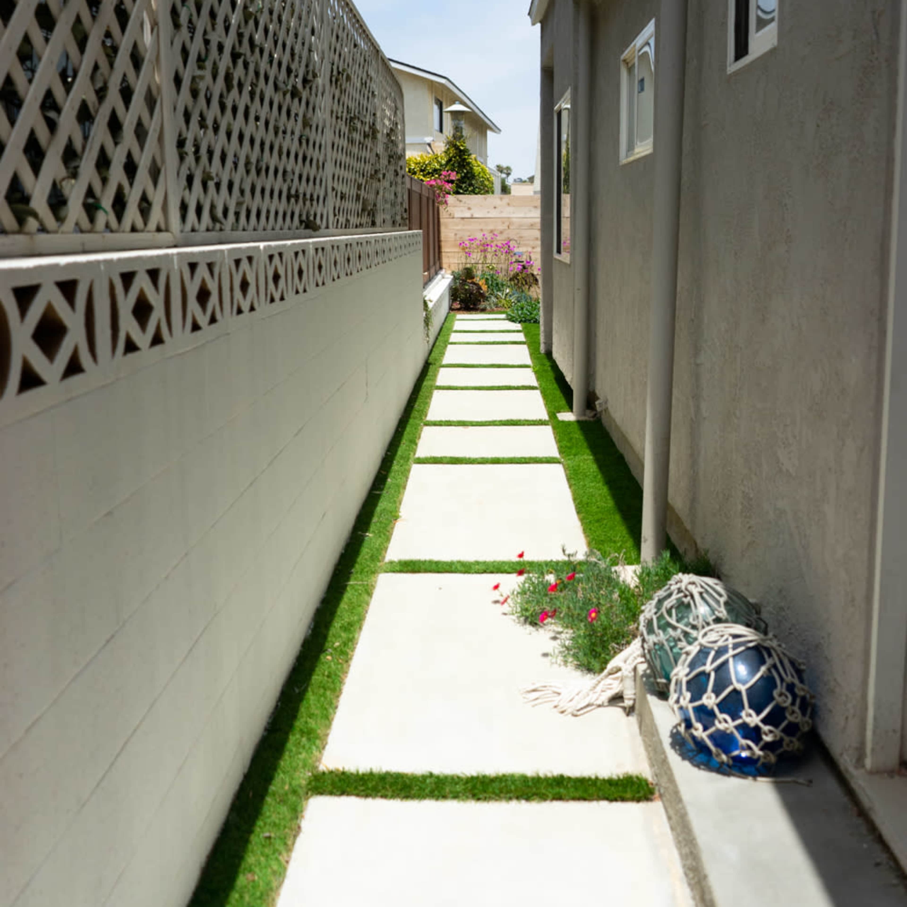 A narrow walkway lined with concrete slabs and patches of artificial grass runs alongside a house, bordered by a lattice fence and flowering plants.