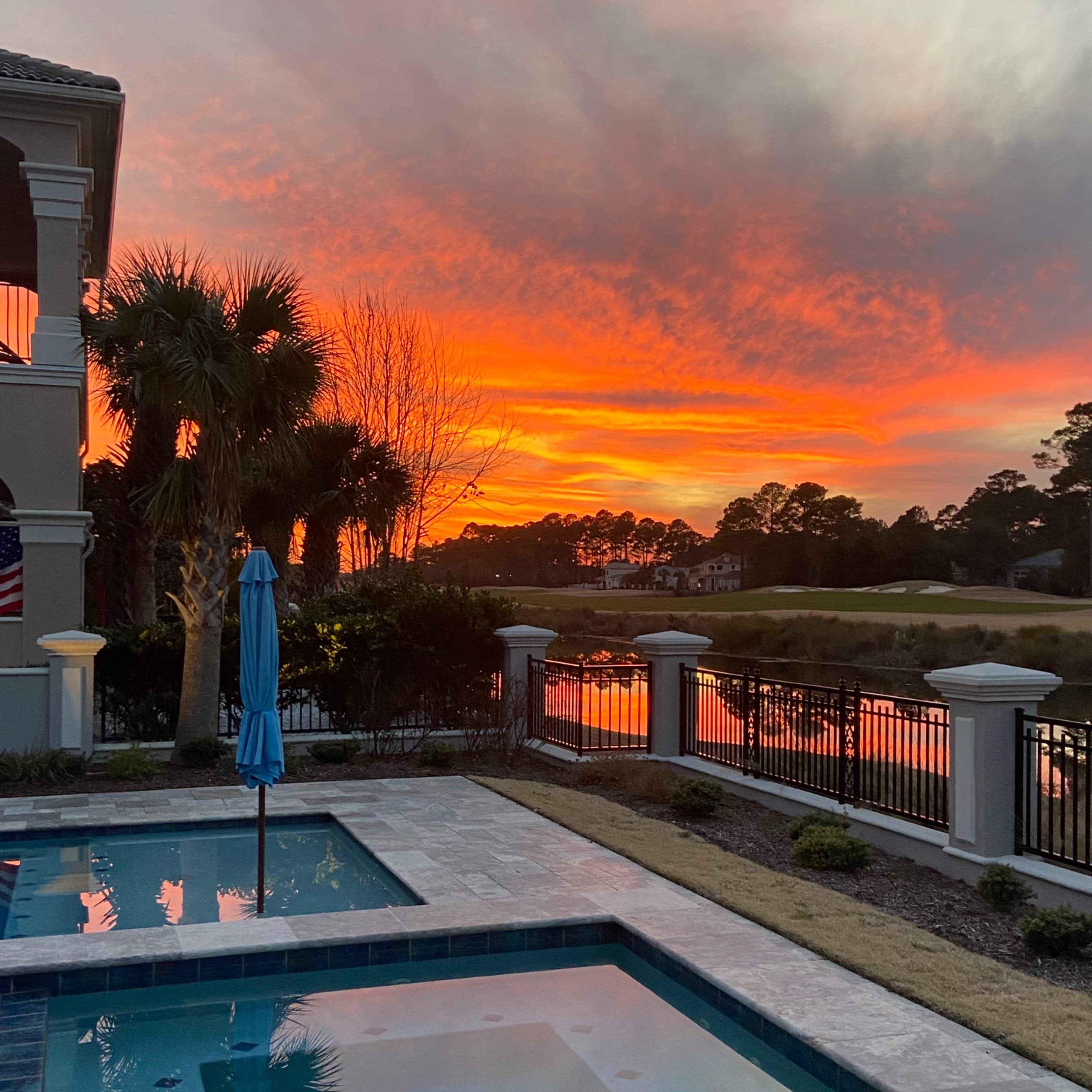 A pool area features a blue umbrella and is framed by palm trees against a vibrant sunset reflecting on the water.