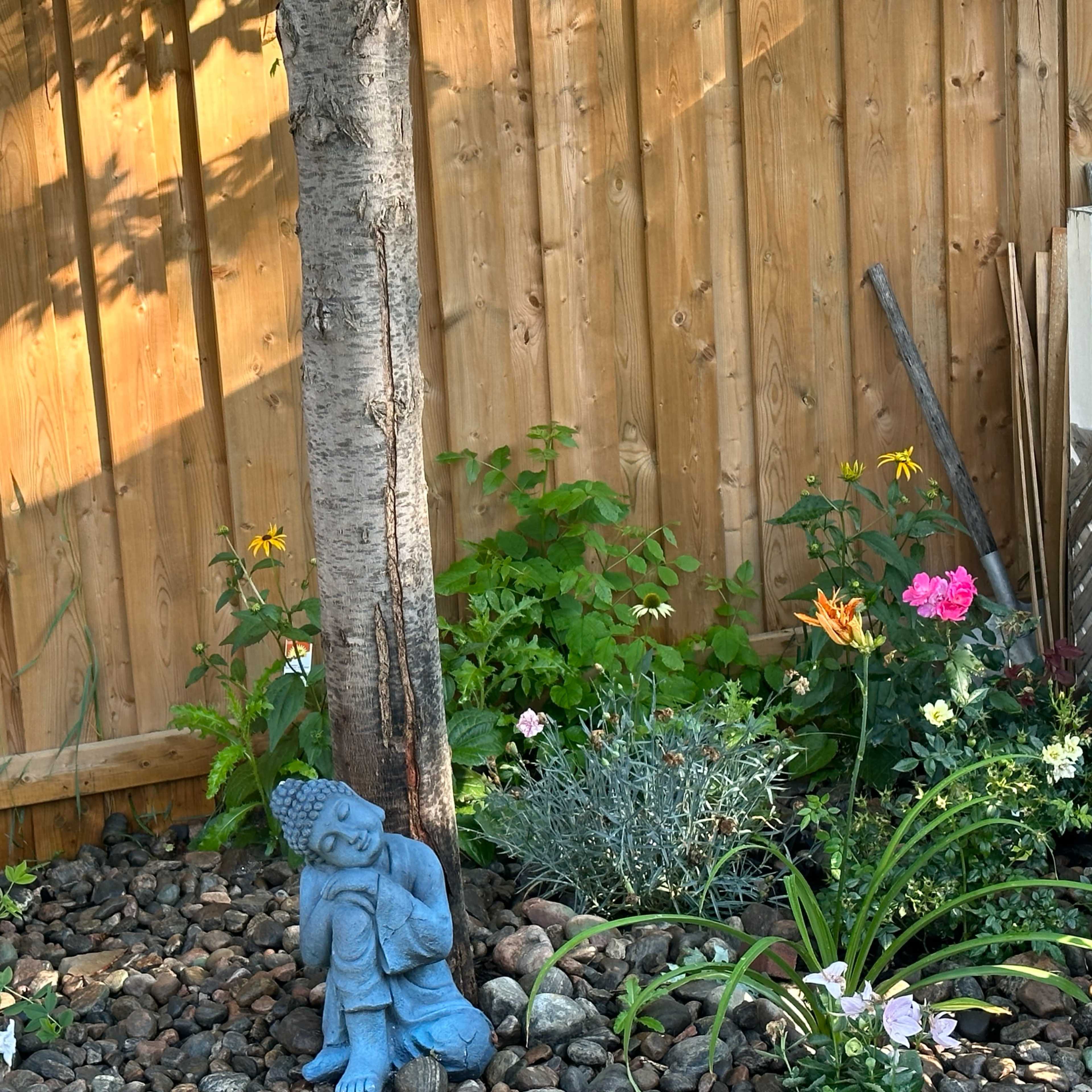 A blue garden statue of a seated figure is placed among various flowers and plants at the base of a tree near a wooden fence.