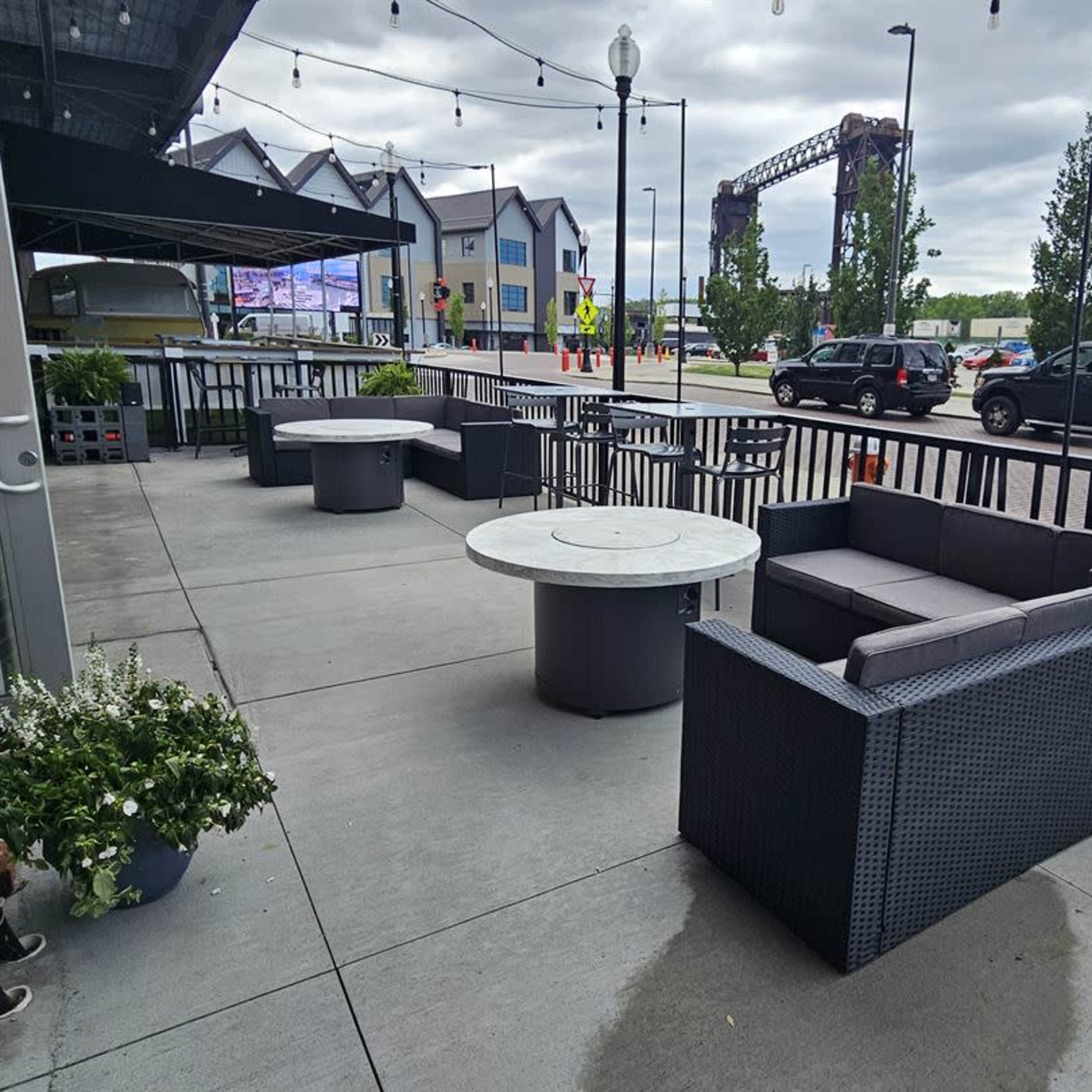 An outdoor seating area with modern furniture, including a round table and upholstered couches, adjacent to a street with visible vehicles and buildings in the background.