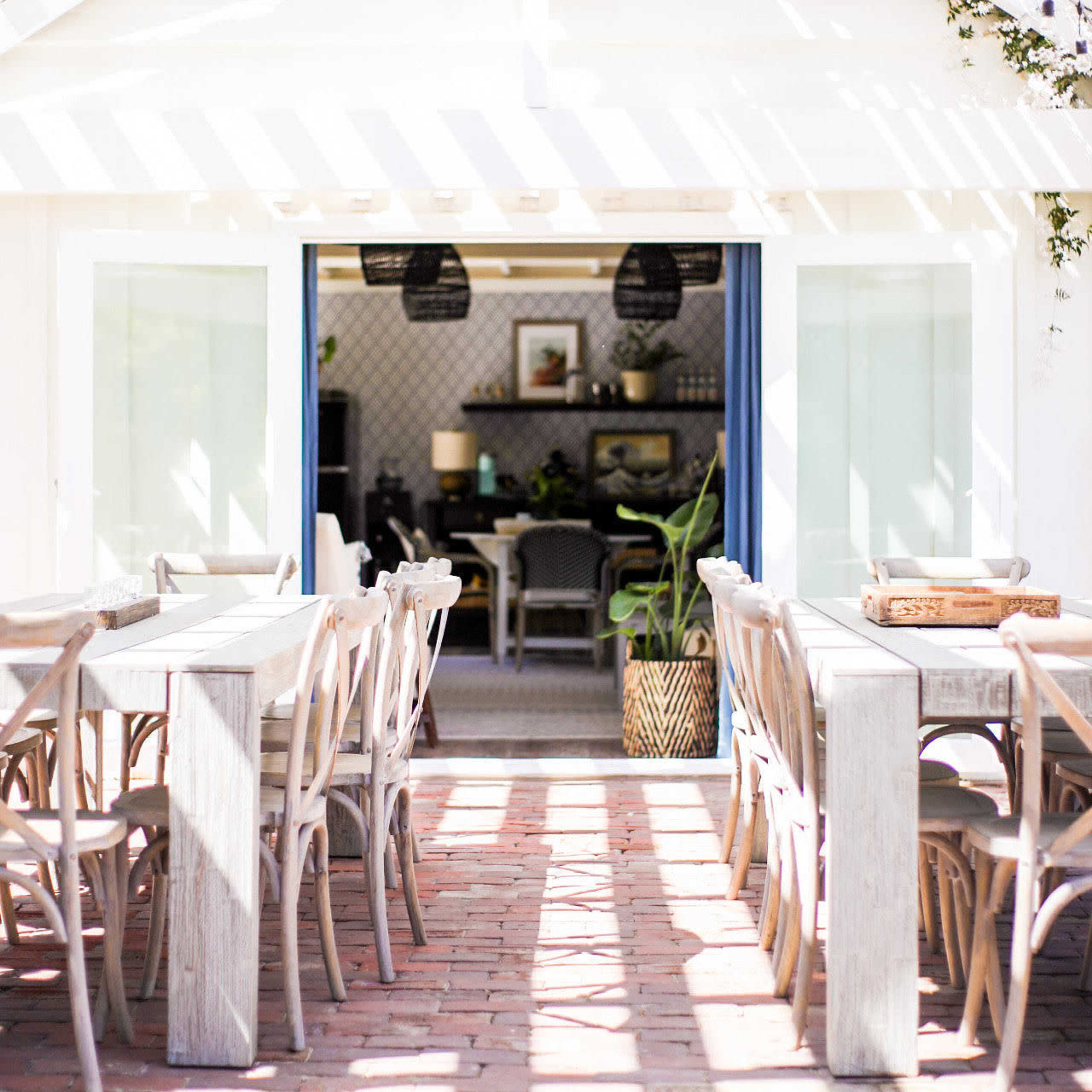 A bright and airy dining area features wooden tables with chairs arranged to face an open entrance leading to a cozy interior space.