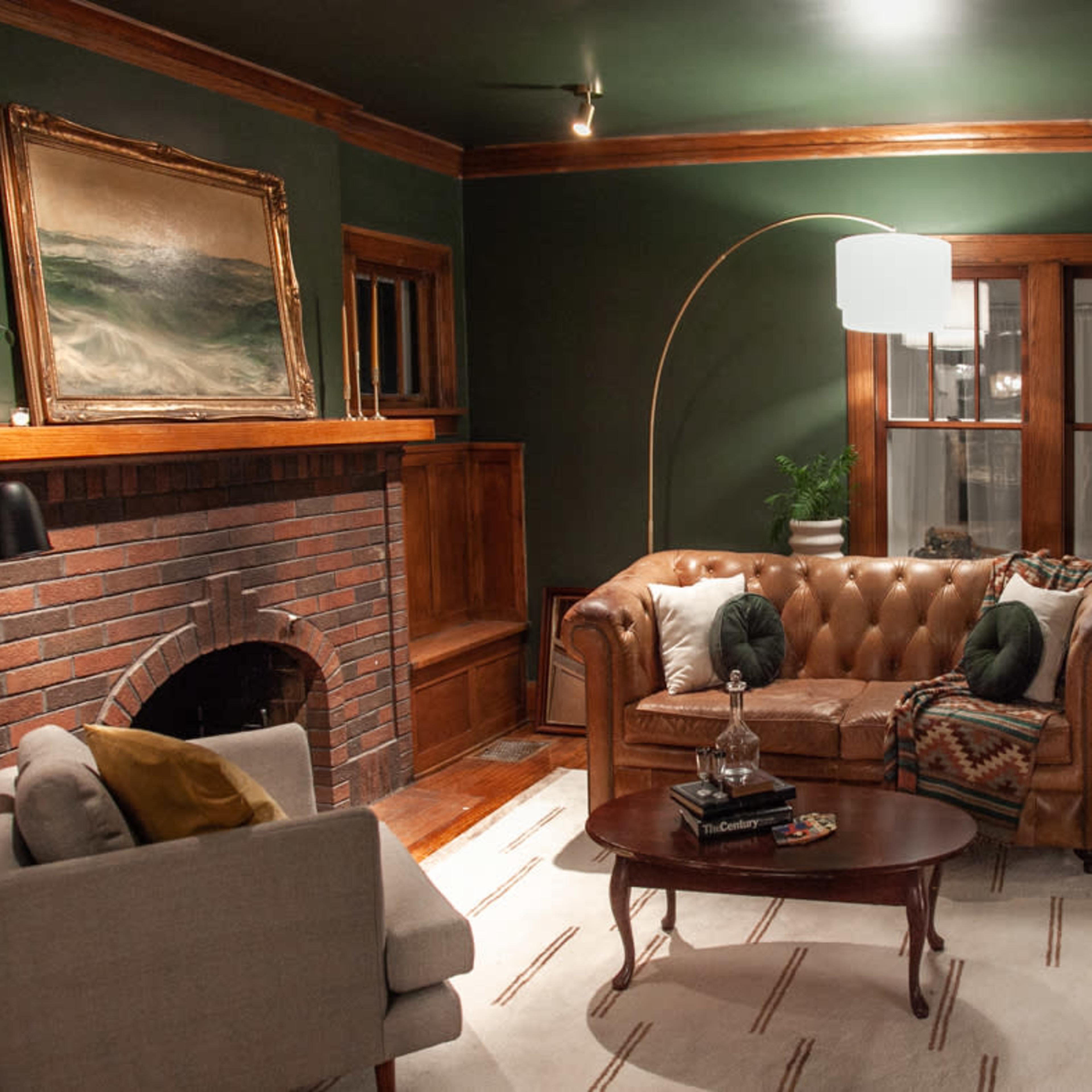 A cozy living room featuring a brown leather chesterfield sofa, a round wooden coffee table, a brick fireplace, and large windows with white curtains.