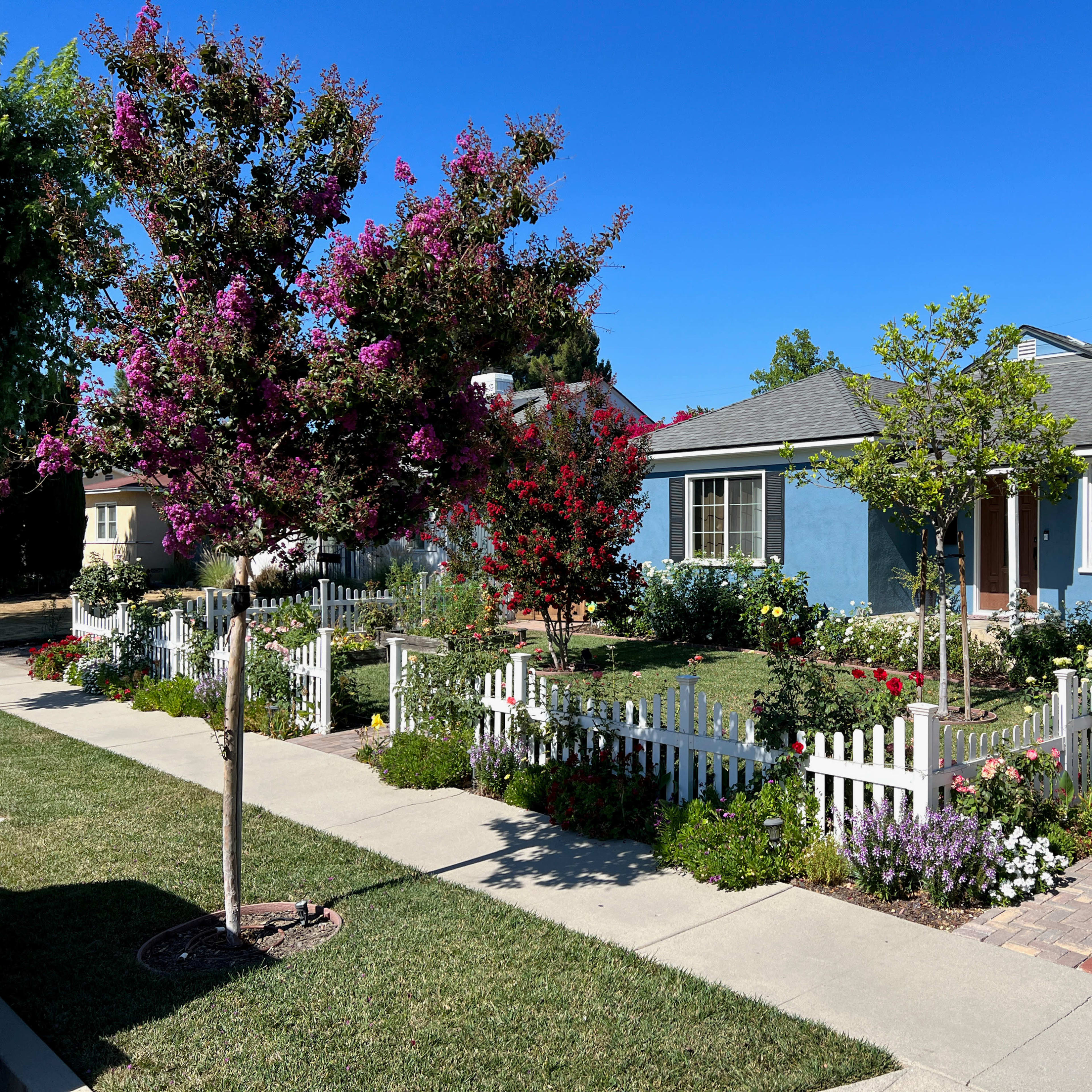 A light blue house with a gray roof is surrounded by colorful flower gardens and a white picket fence.